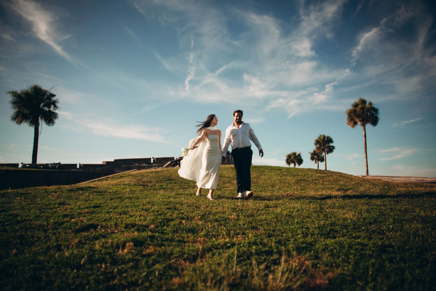 Bride and groom holding hands running on green hill with palm trees and blue sky at outdoor wedding venue.