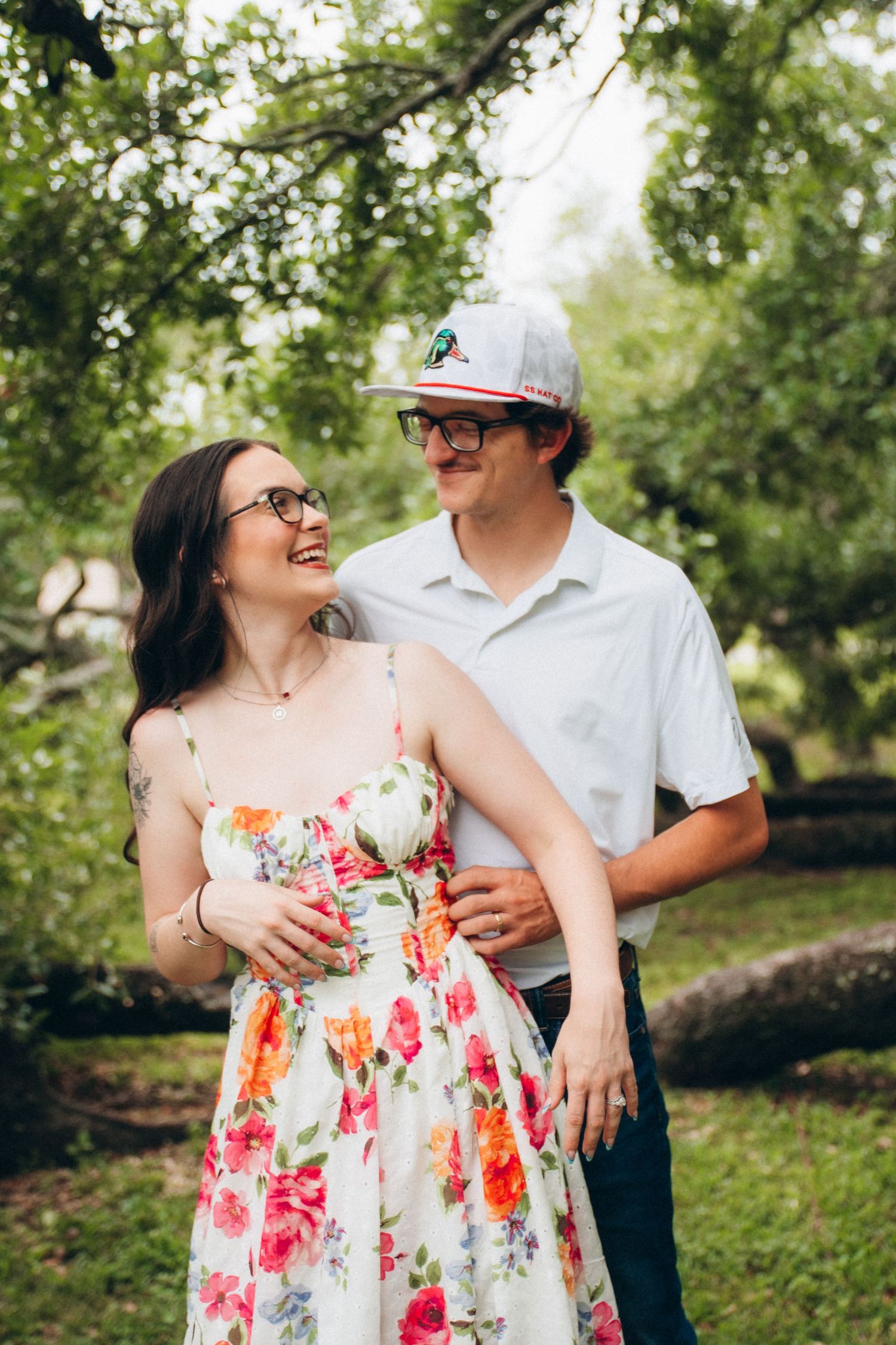 Laughing couple embracing in green park, woman in colorful floral sundress with tattoos, man in white shirt and cap.