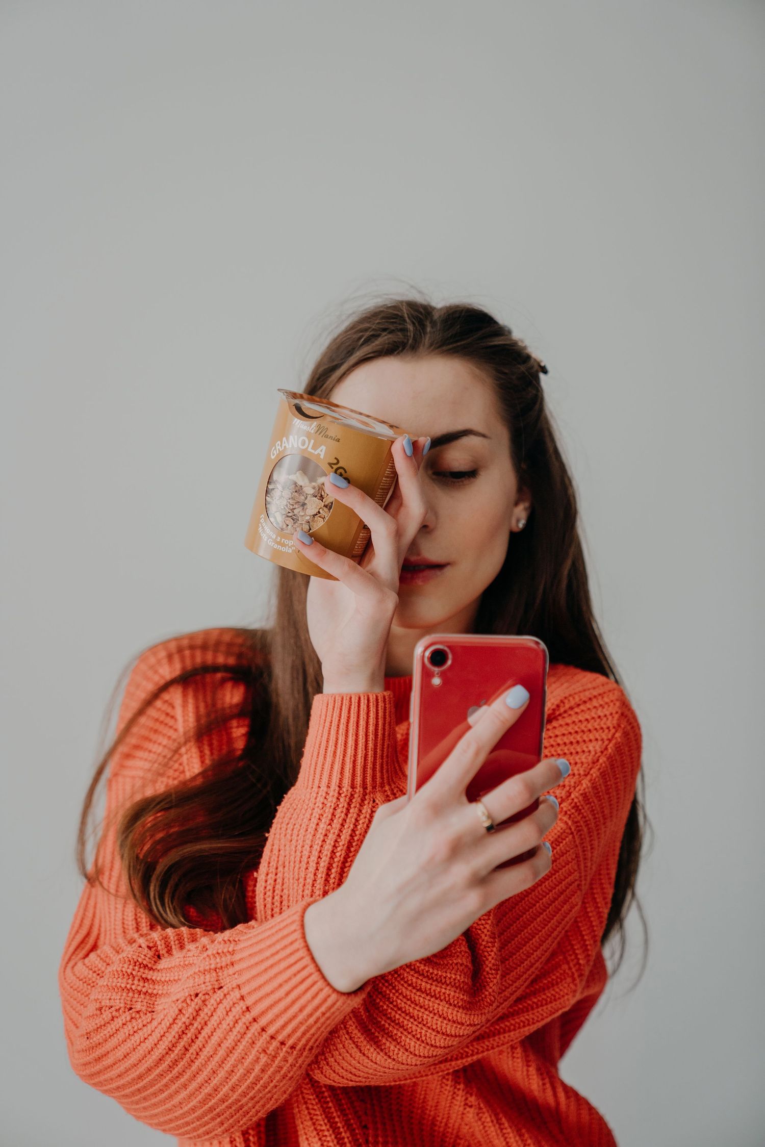 Woman in orange sweater holding a small bag over her face while looking at her red smartphone.