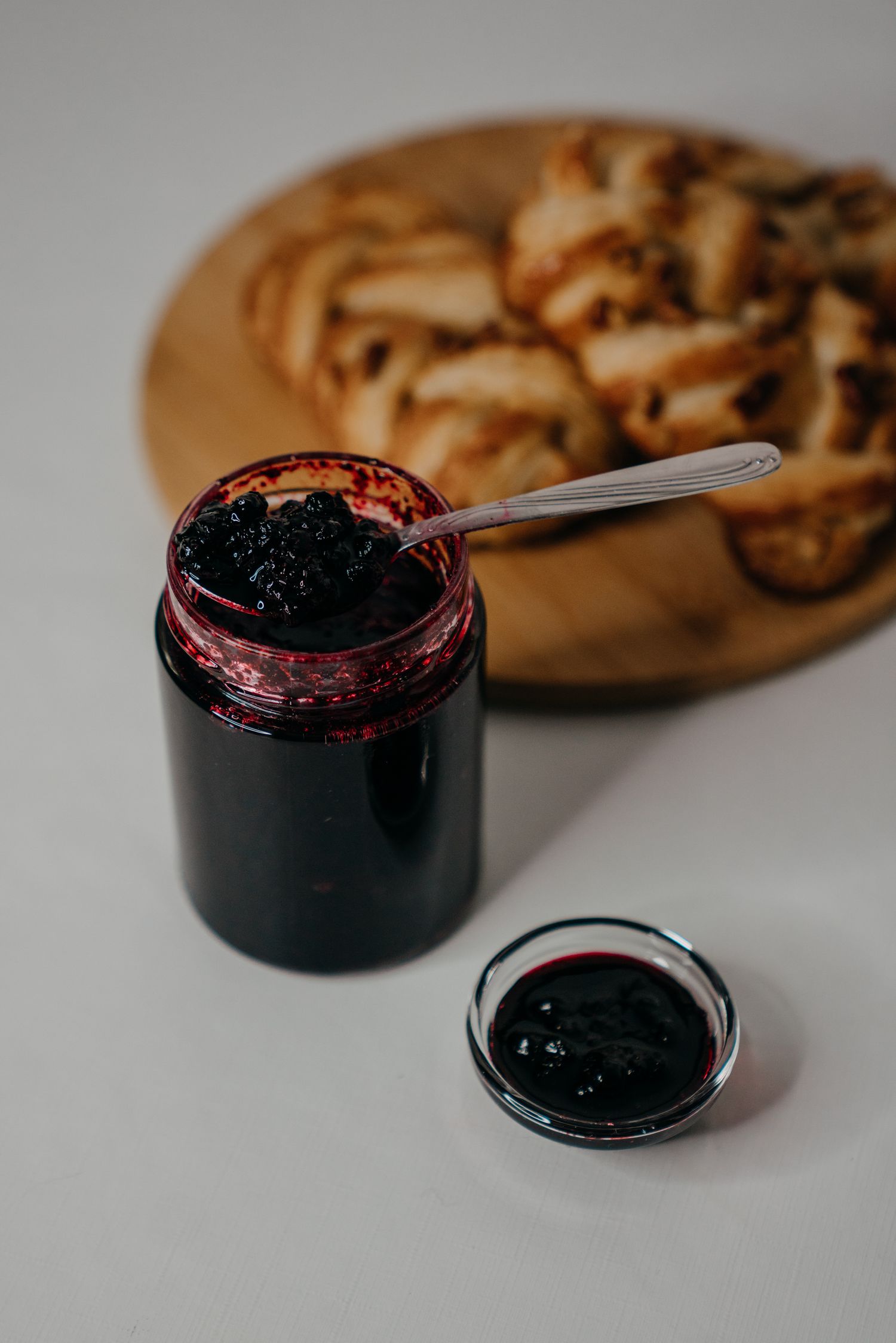 Dark berry jam in an open glass jar with a spoon, beside a small dish, with pastries on a wooden board in background.