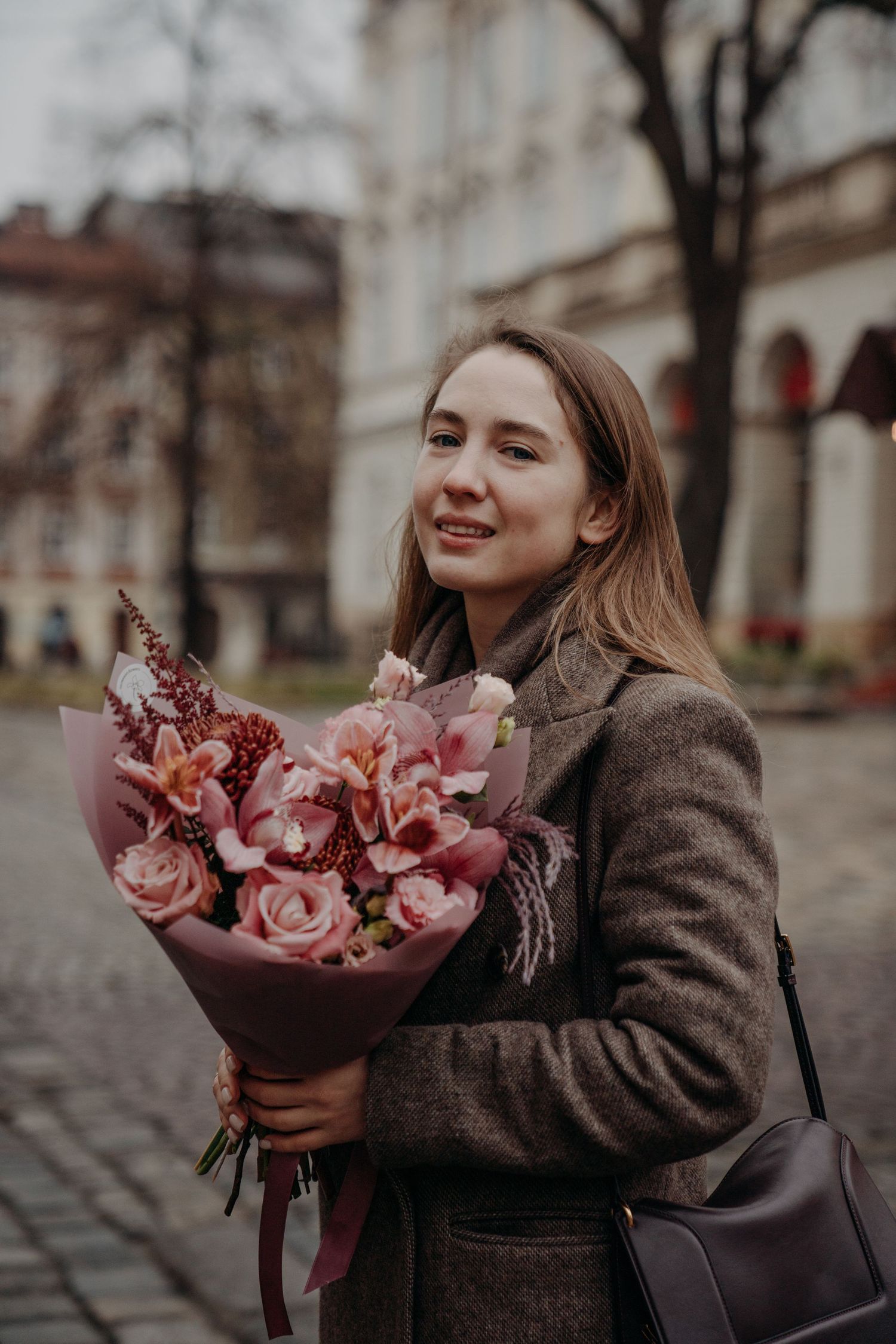Smiling woman in gray coat holding pink floral bouquet on a cobblestone city square.