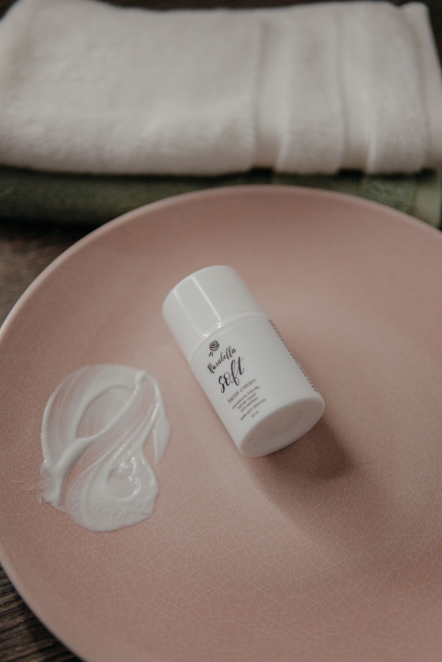 White skincare moisturizer stick displayed on a pink ceramic plate with cream swatch and folded towels in background.