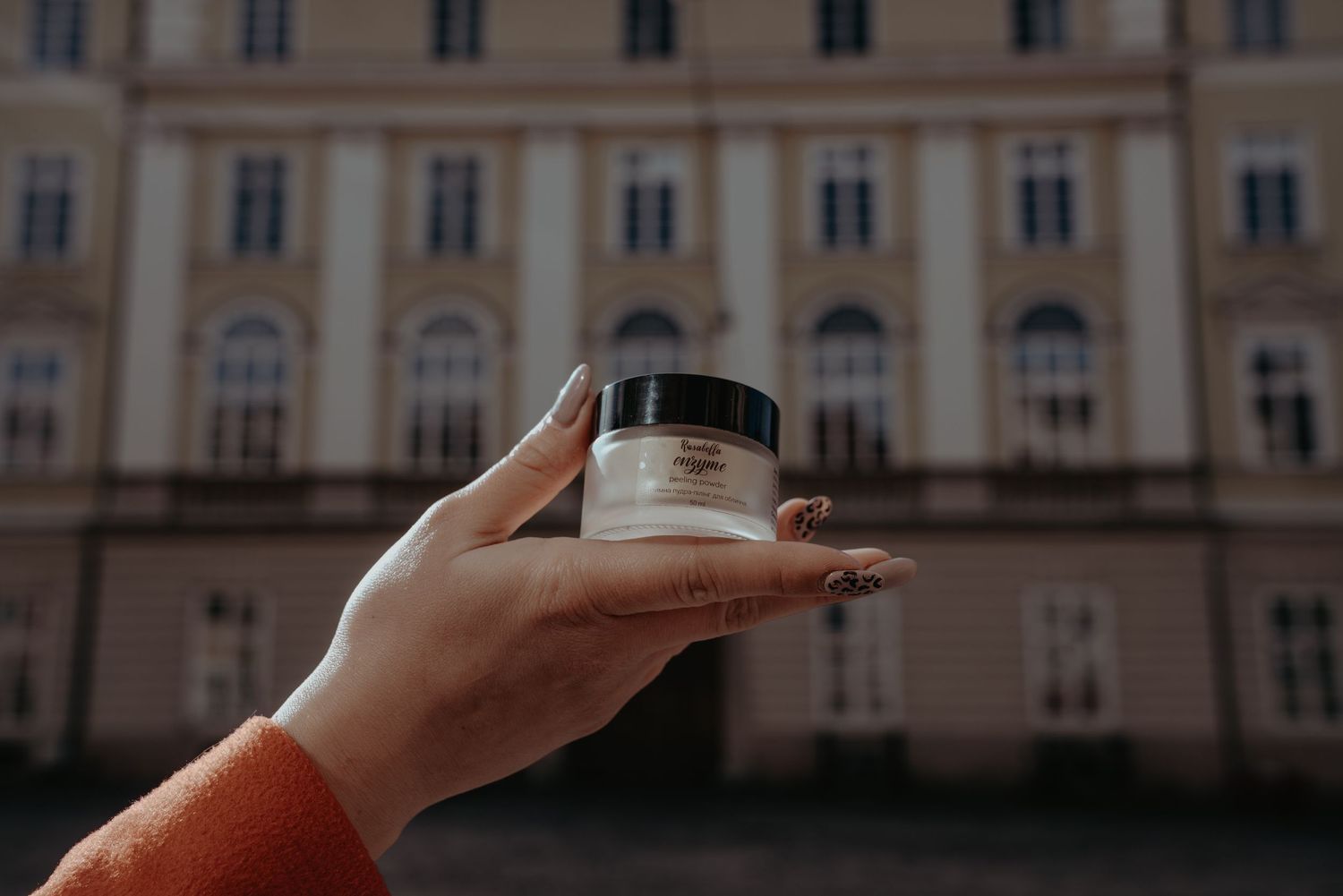 Hand holding a small white cosmetic jar in front of a classic European building facade.