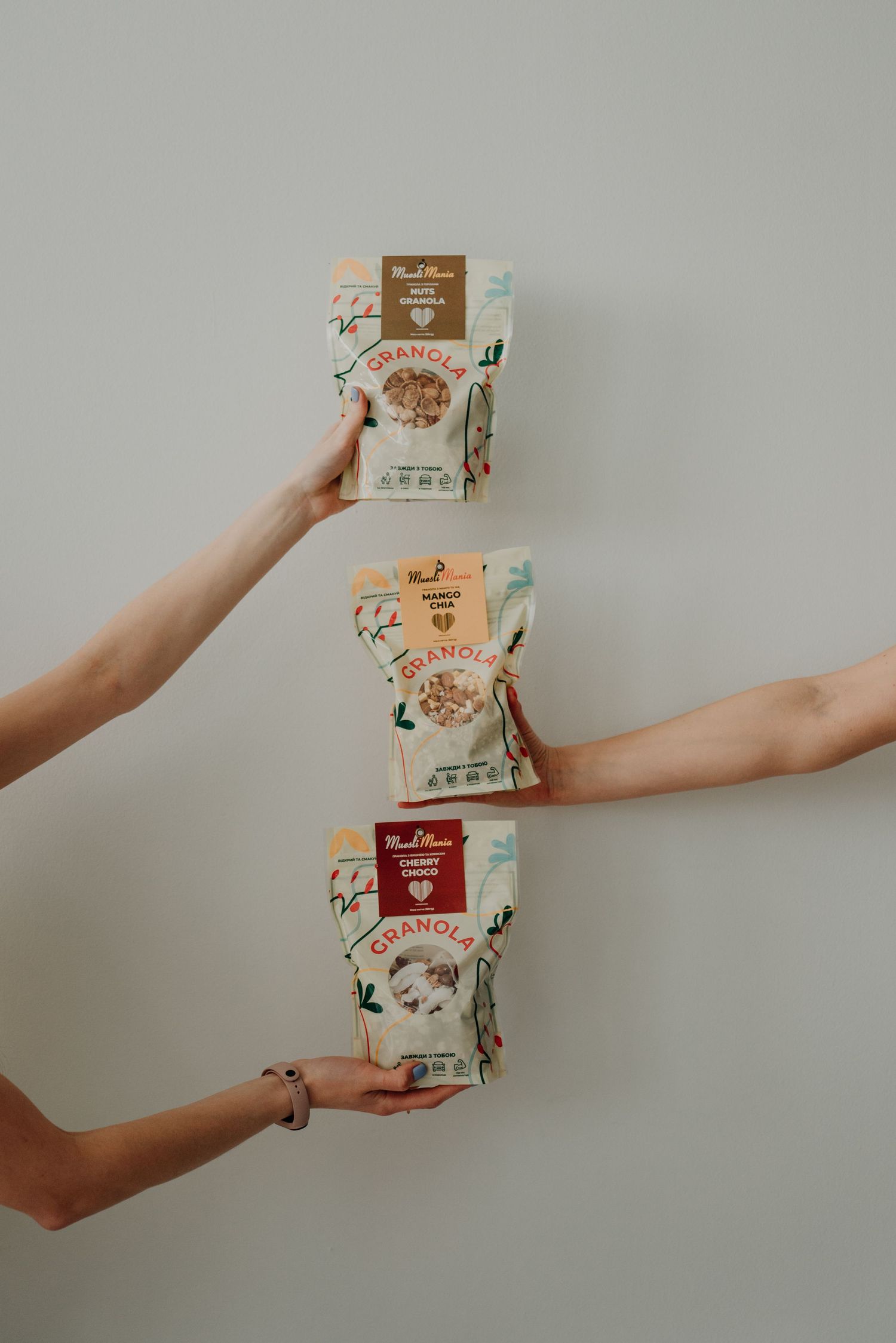Three hands holding up different flavored granola packaging bags against a light gray background.