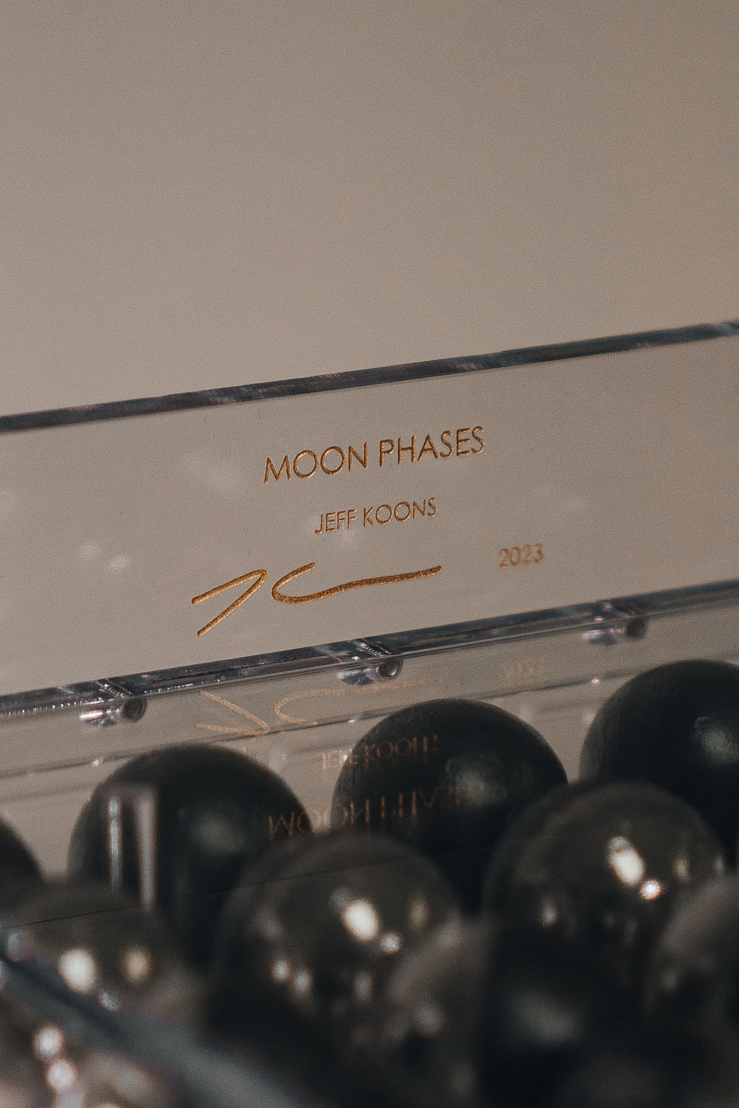 Close-up of Jeff Koons Moon Phases artwork label with gold signature dated 2023 and dark spherical objects below.