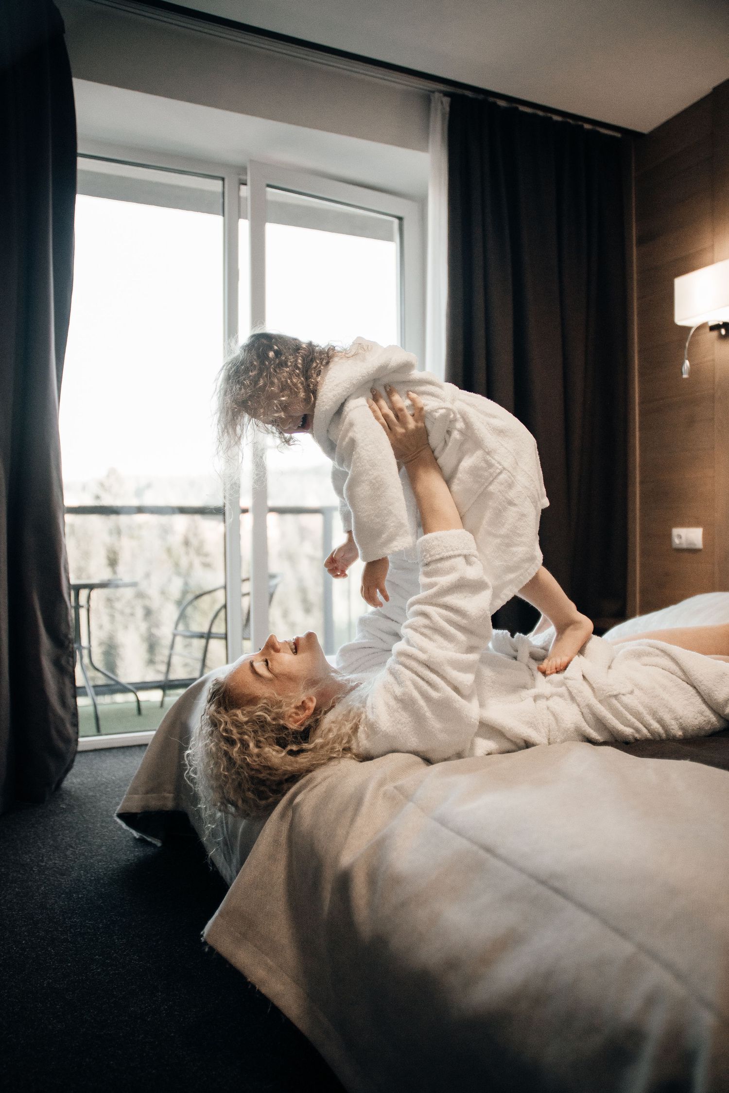 Mother in white bathrobe lying on bed lifting her young child up in a bright hotel room with large window.