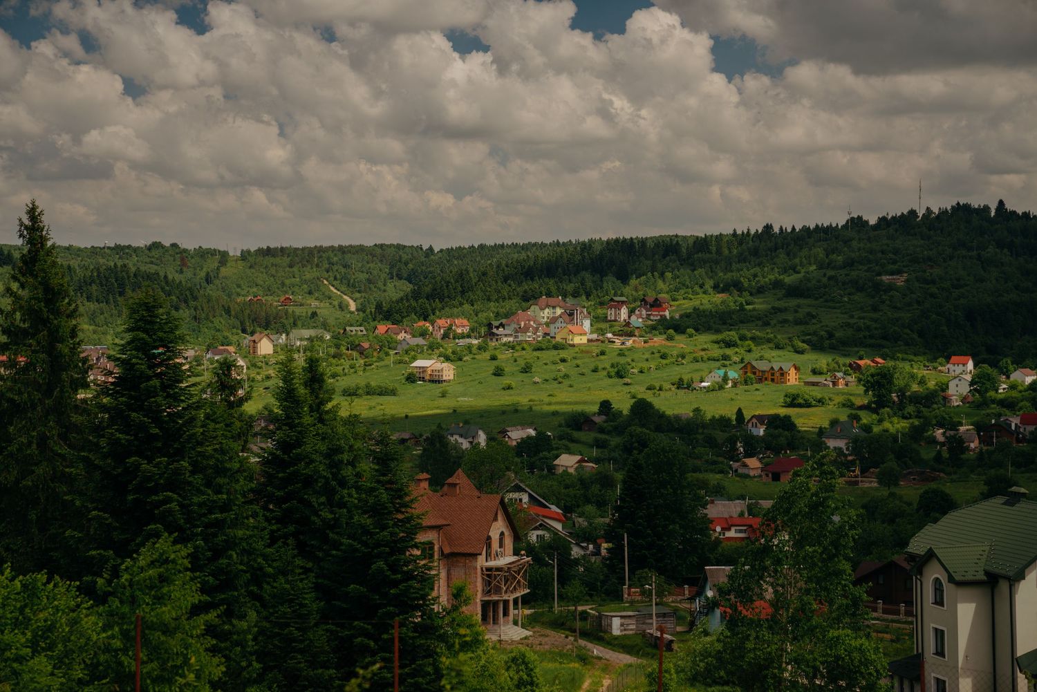 Scenic mountain village with colorful houses nestled among lush green hills and pine trees under dramatic cloudy sky.