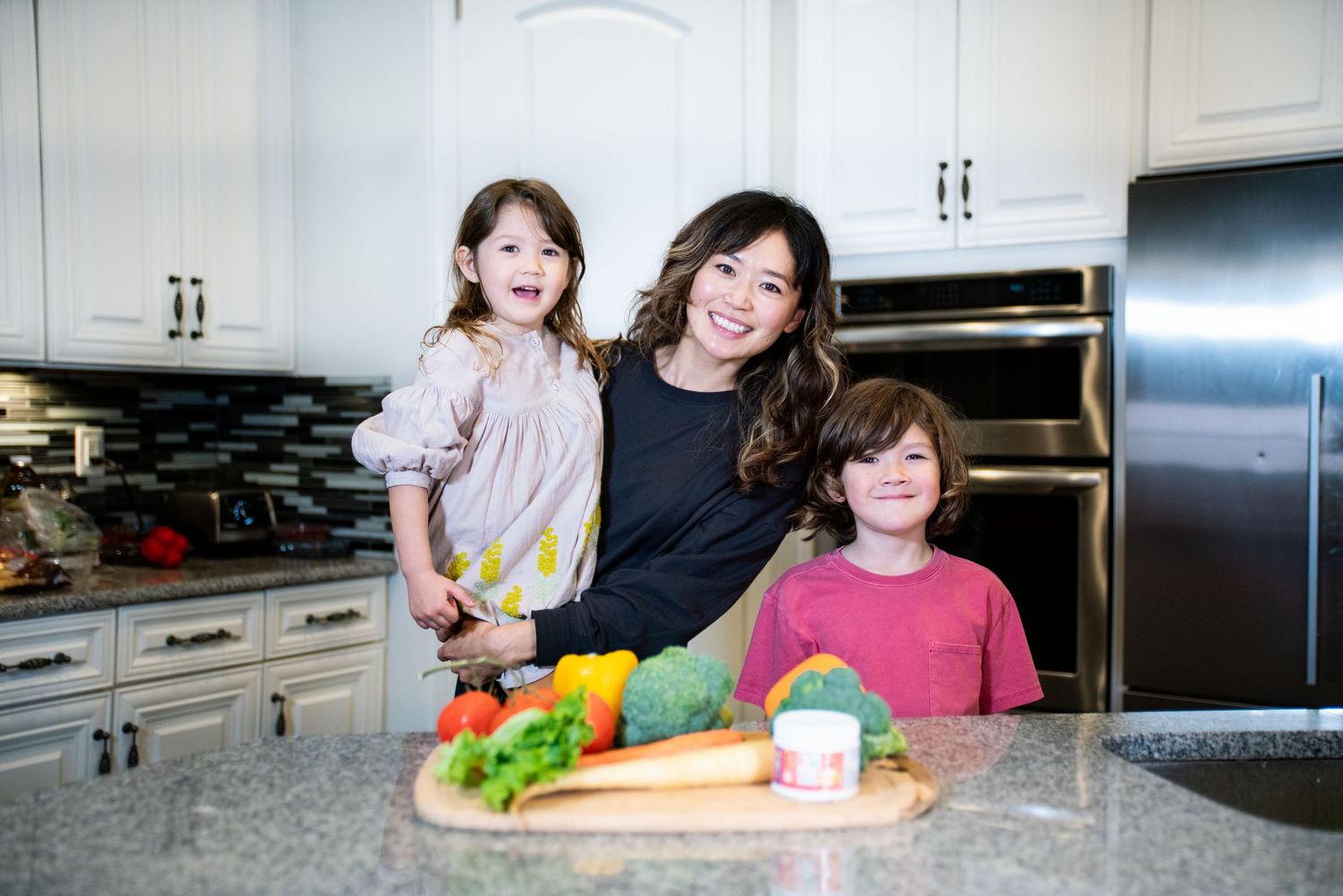 Mom and two kids smiling in kitchen with fresh vegetables on cutting board.