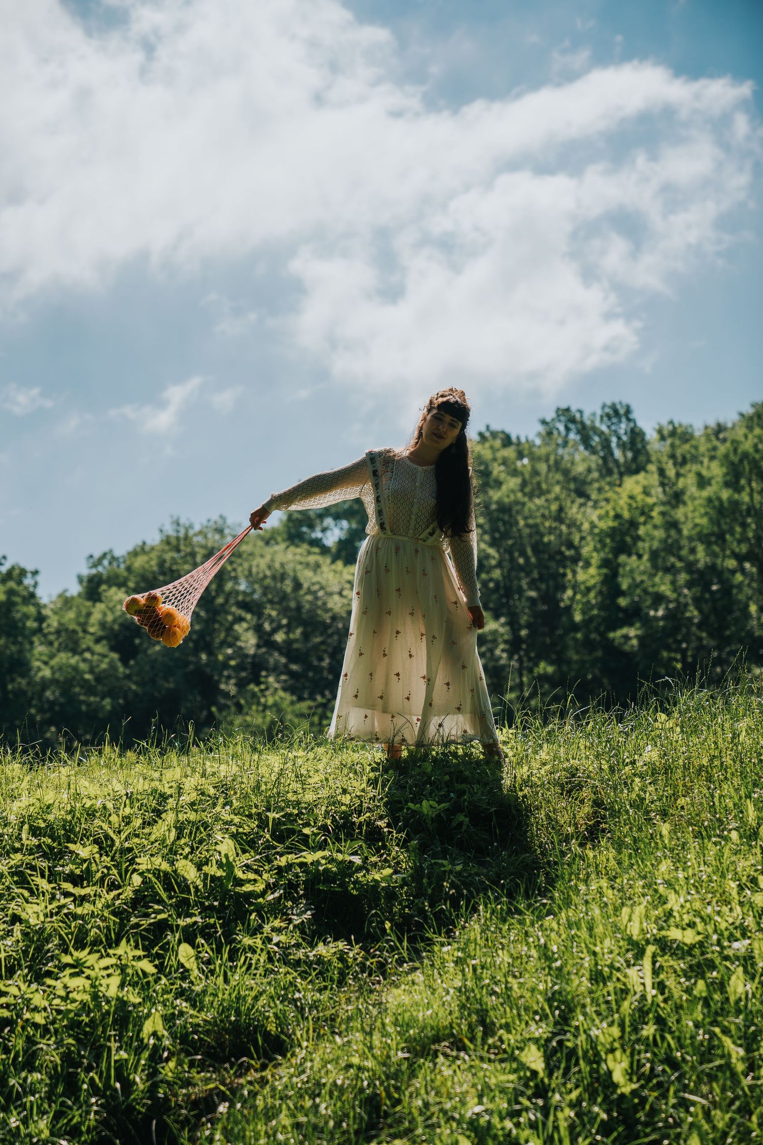 Woman in white floral dress holding a mesh bag with flowers, standing on a green hillside under a blue sky.