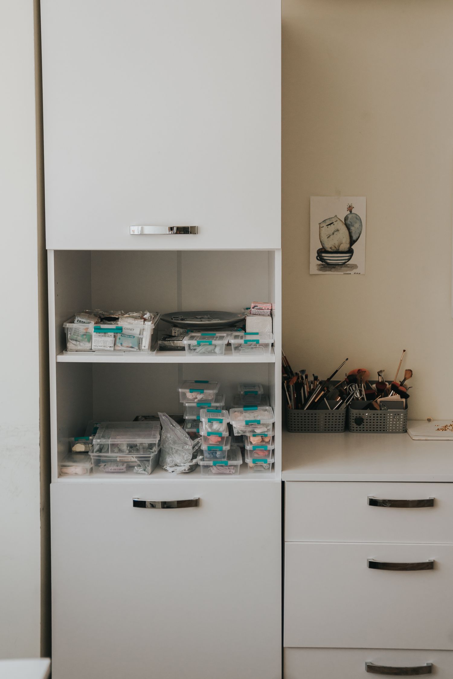 White cabinet shelving unit storing craft supplies and materials next to a desk with tools in a studio.