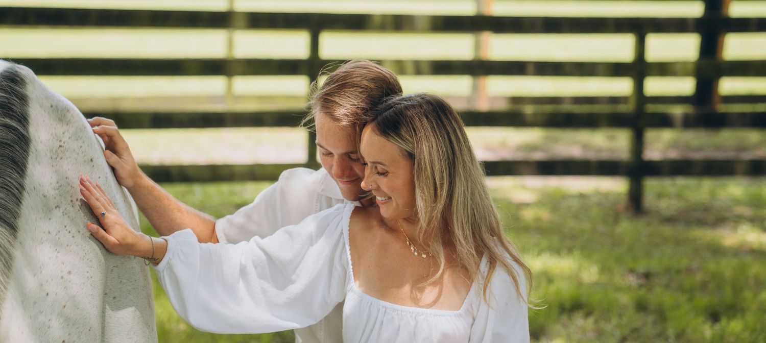 A couple and their white horse share tender moments in a scenic pasture setting.