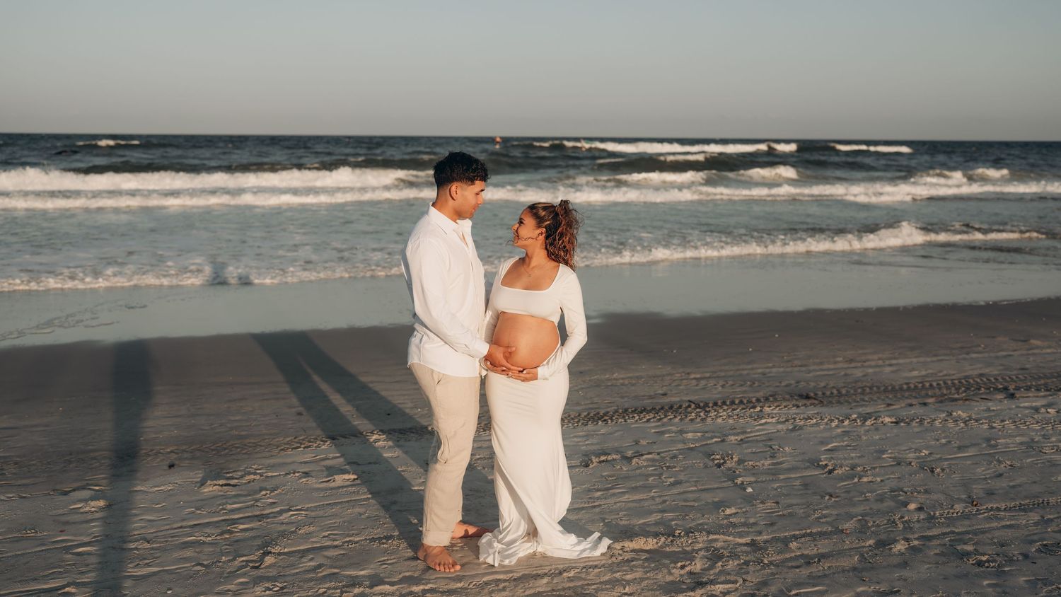 Panoramic beach scene with a couple taking maternity photos at the water's edge.