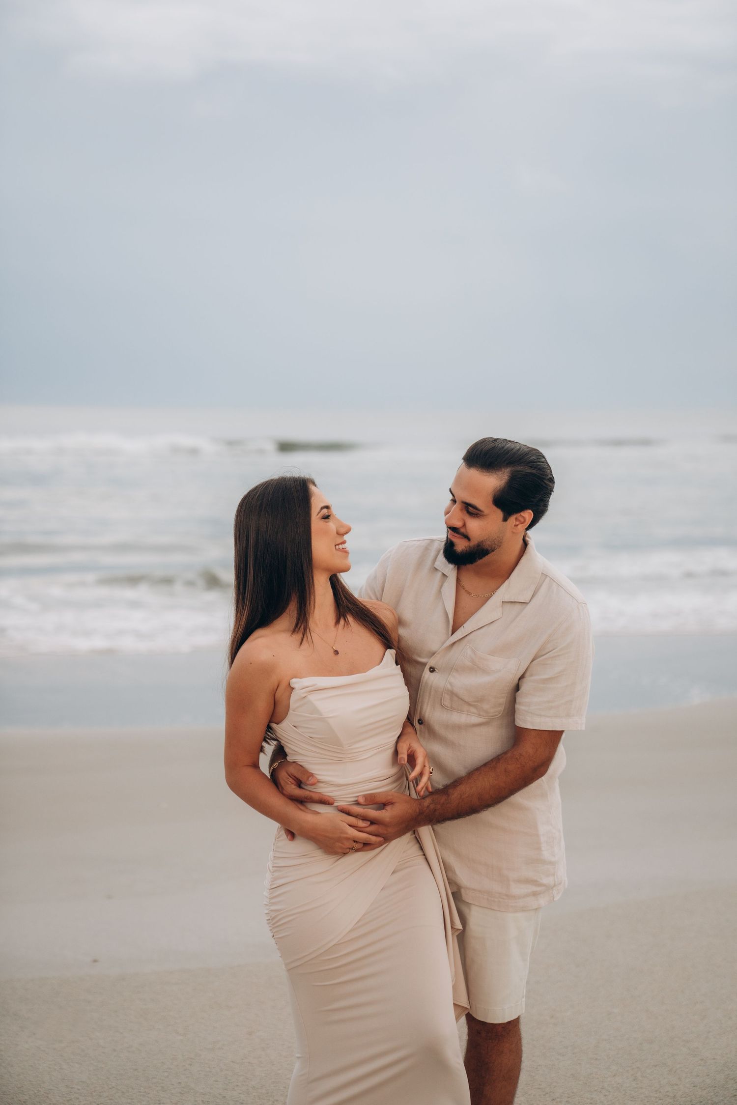Couple in light colored outfits sharing tender moment on beach with cloudy sky.