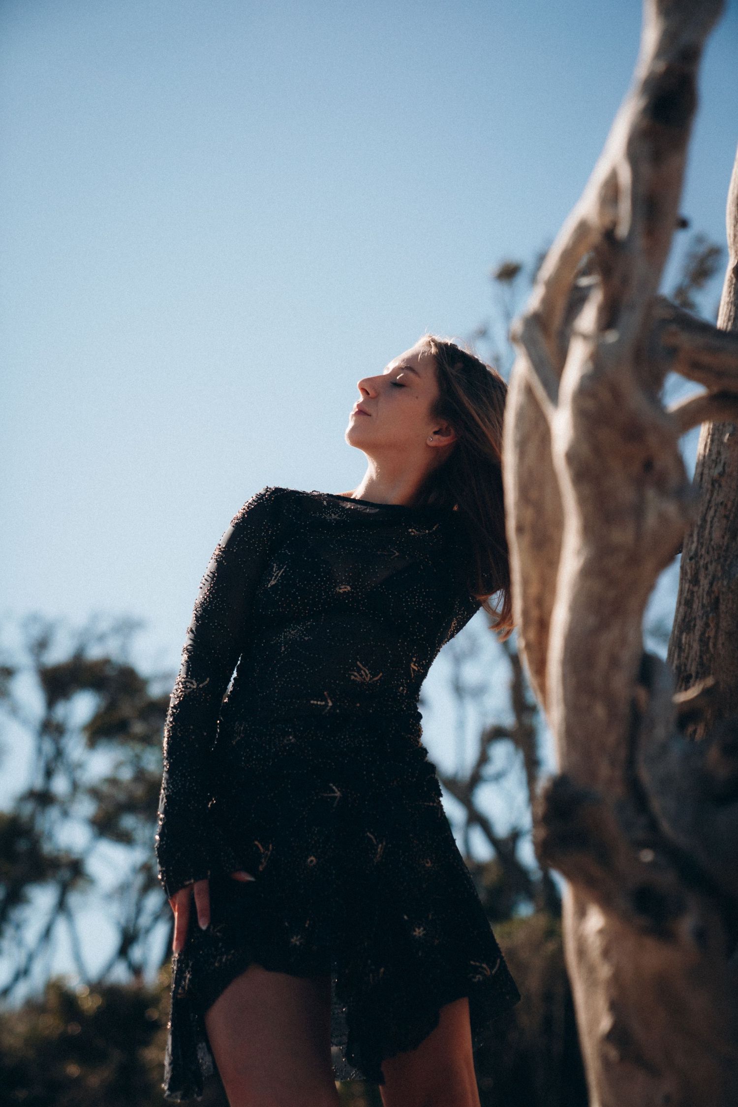 A person in a black outfit poses against a bright sky with tree branches in the background.