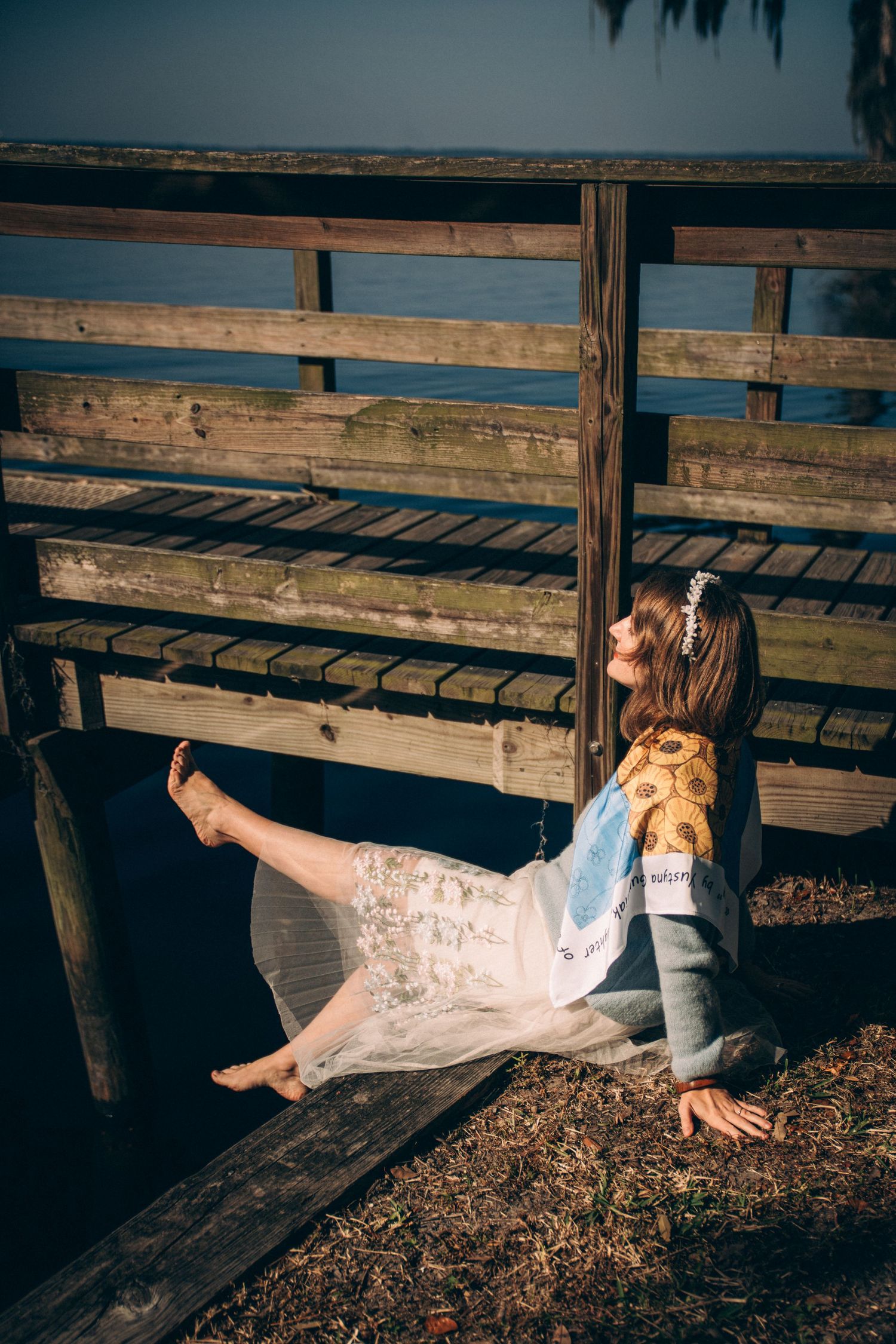 A person in white dress lying on wooden boardwalk planks near a waterfront at sunset.