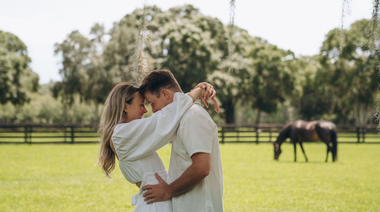 Couple in white outfits share an intimate moment in a pastoral horse farm setting.