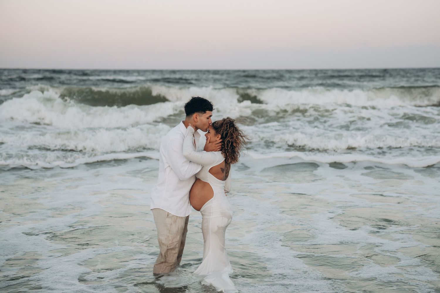 A couple in white clothing plays in the ocean waves during a beach photoshoot.