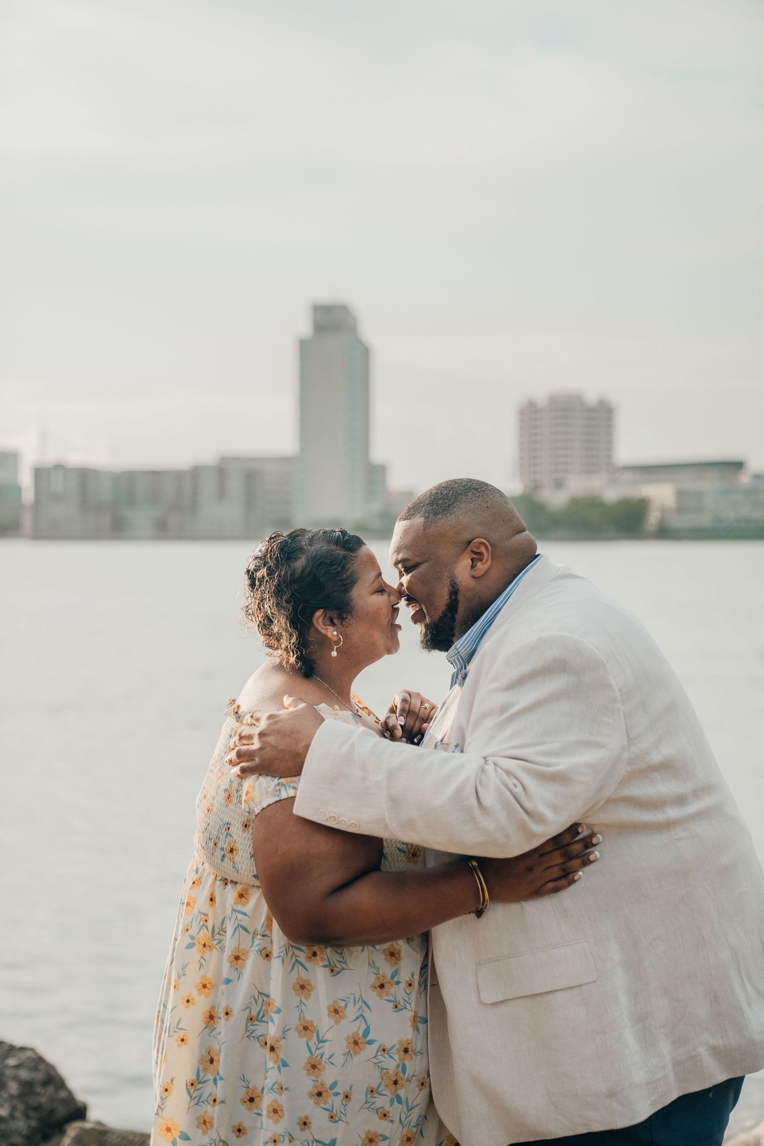 A couple embraces by the waterfront with a city skyline in the background.