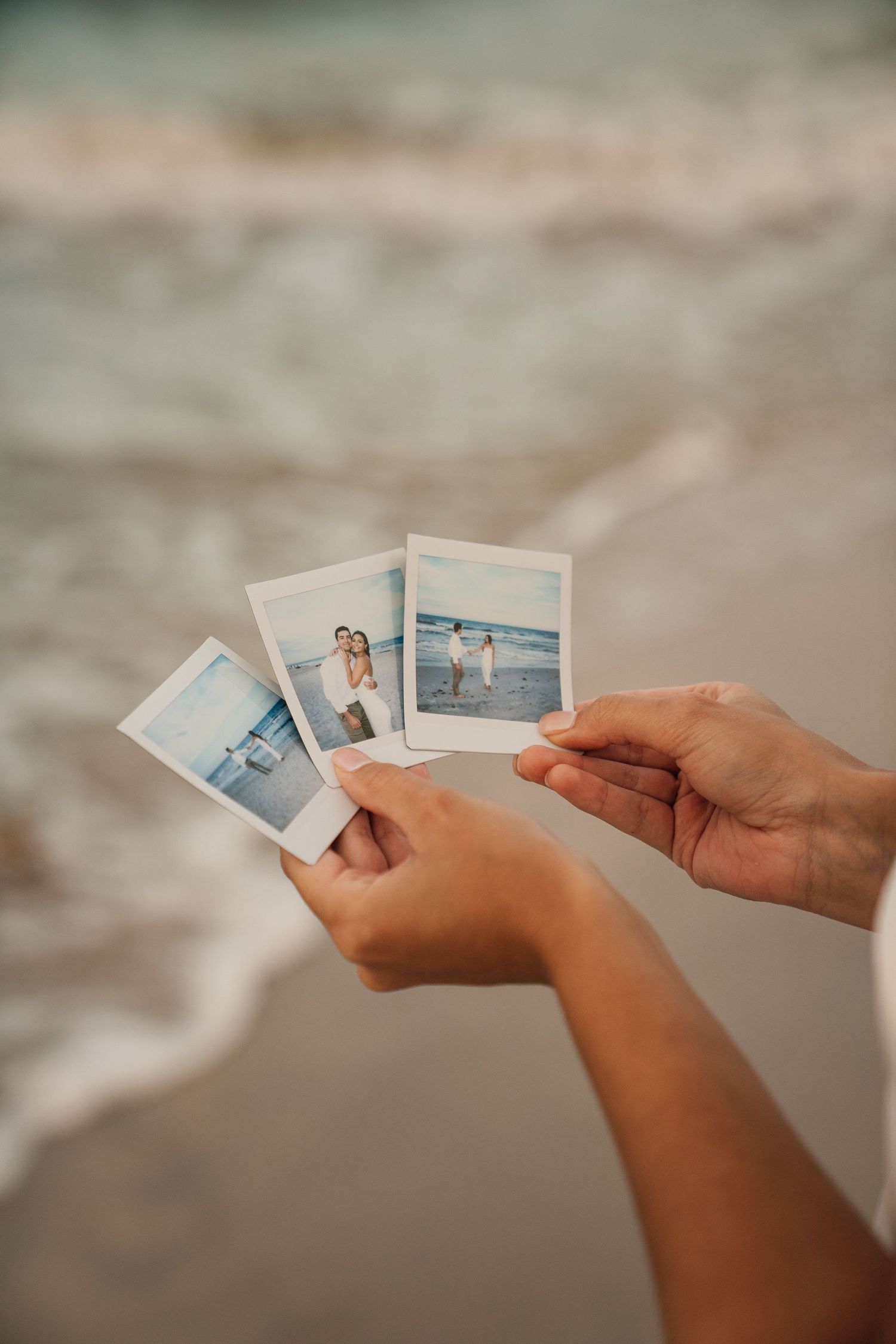 Hands holding instant photos against a sandy beach background with ocean waves.