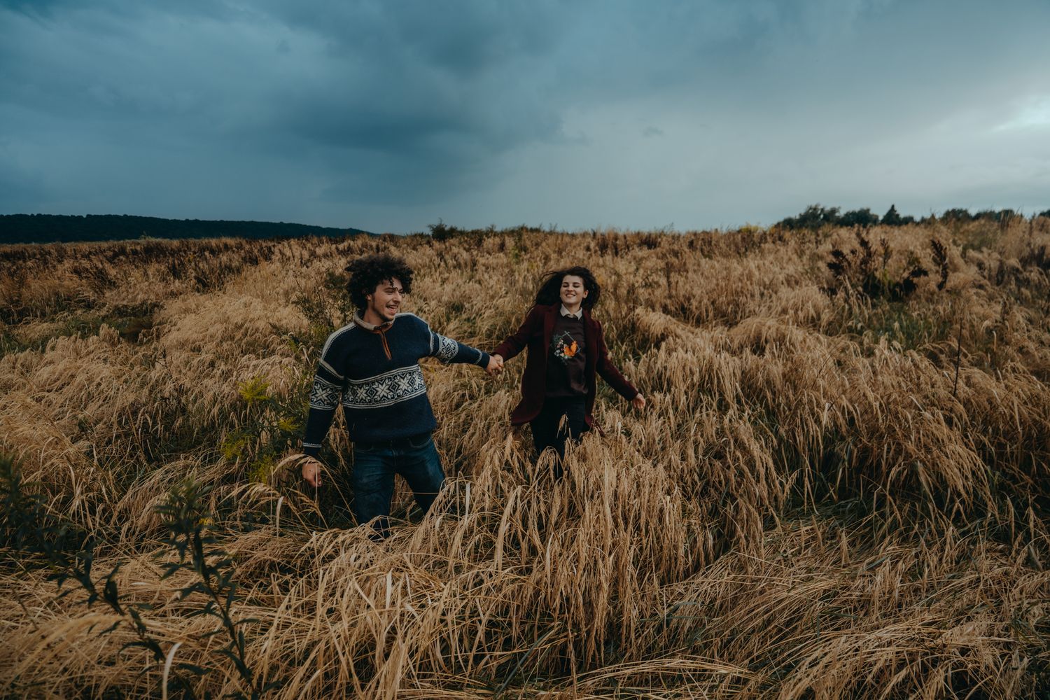 Playful couple running through dry autumn grass under moody skies.