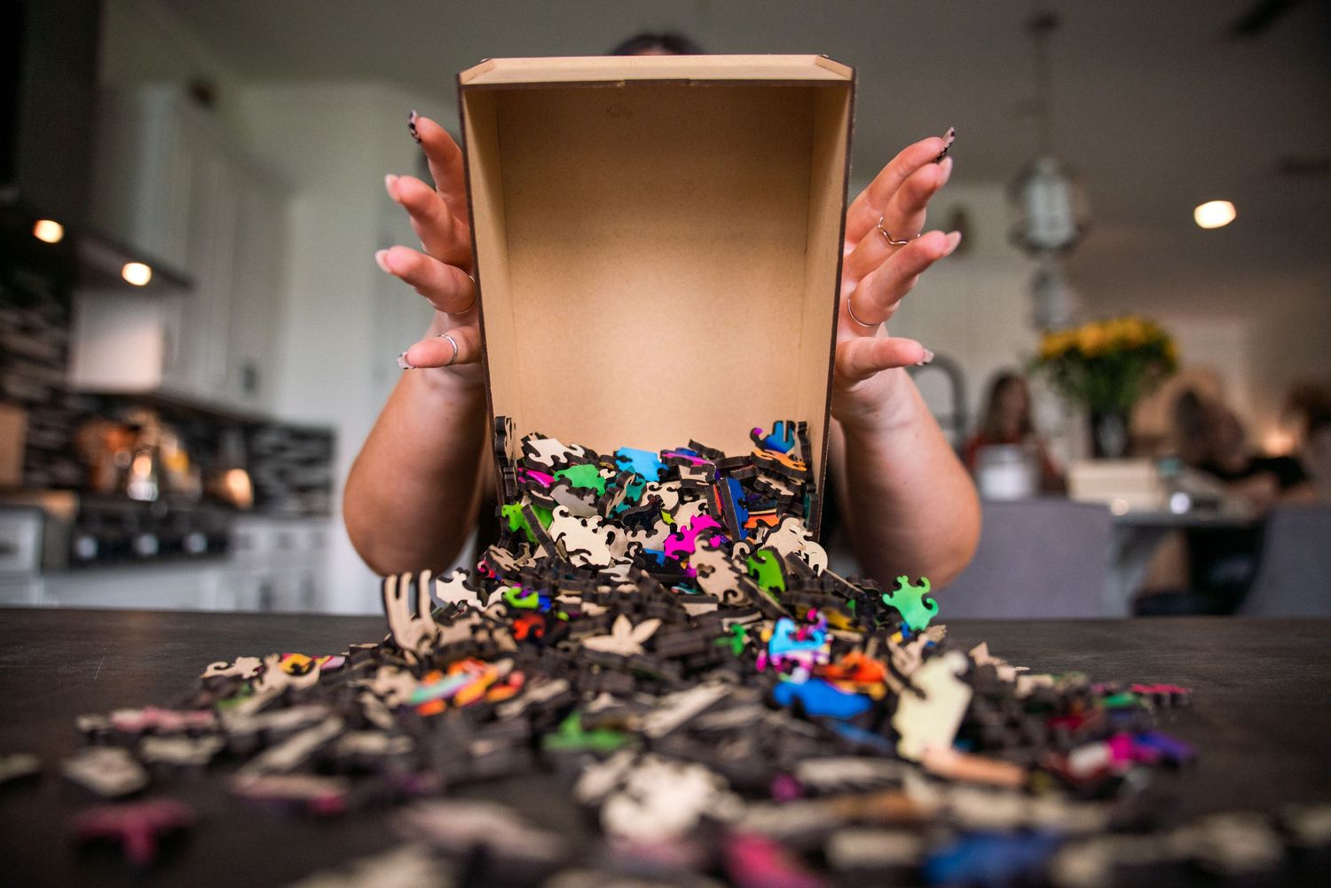 Hands pouring colorful wooden puzzle pieces from a cardboard box onto a wooden table.