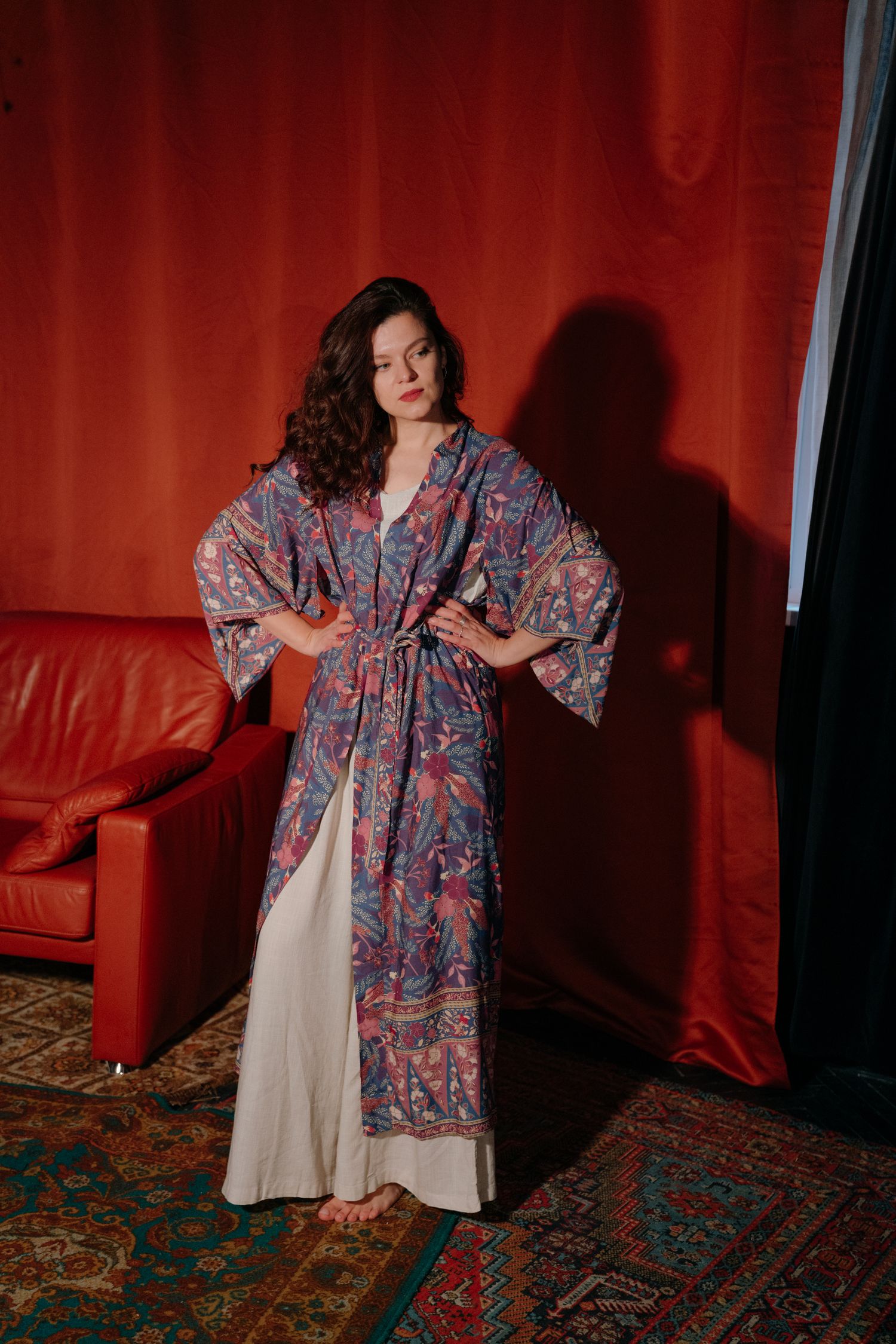 Woman in floral kimono robe over white dress poses confidently against red curtain backdrop on patterned rug.
