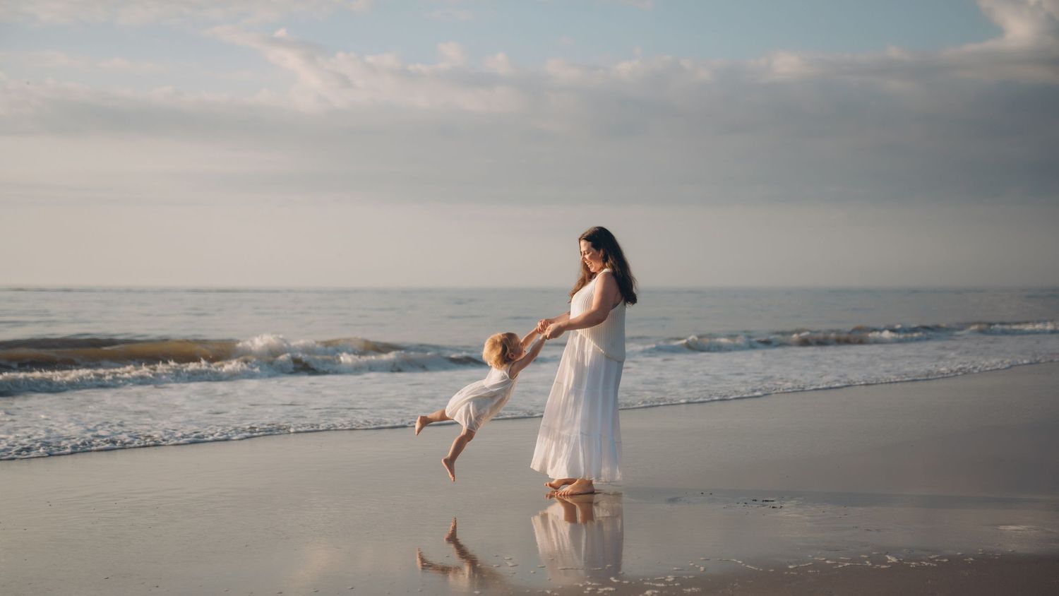 A mother in white dress joyfully swings her toddler on a serene beach with ocean waves in the background.