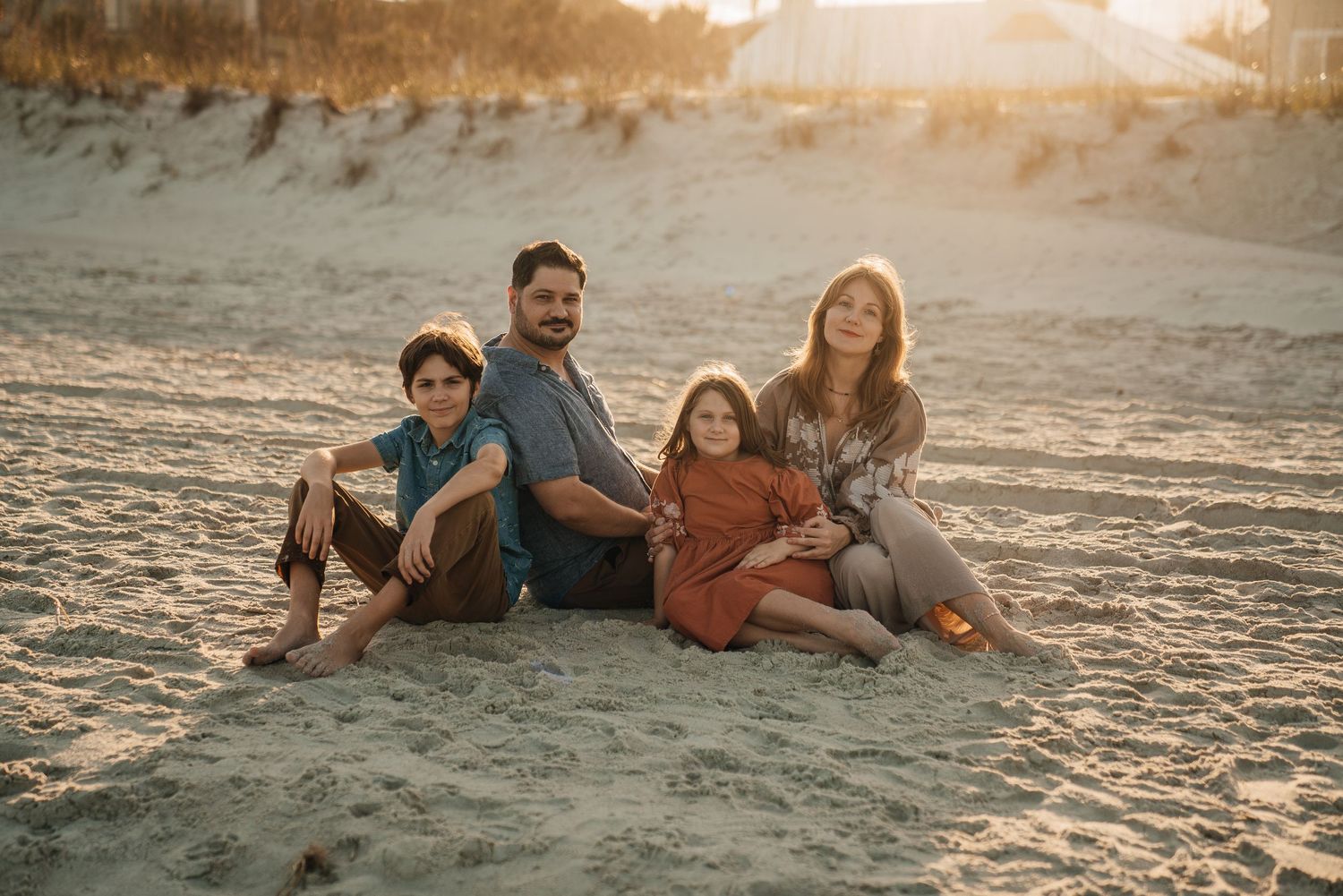 Family of four sitting together on sandy beach at golden hour sunset, smiling warmly at camera.