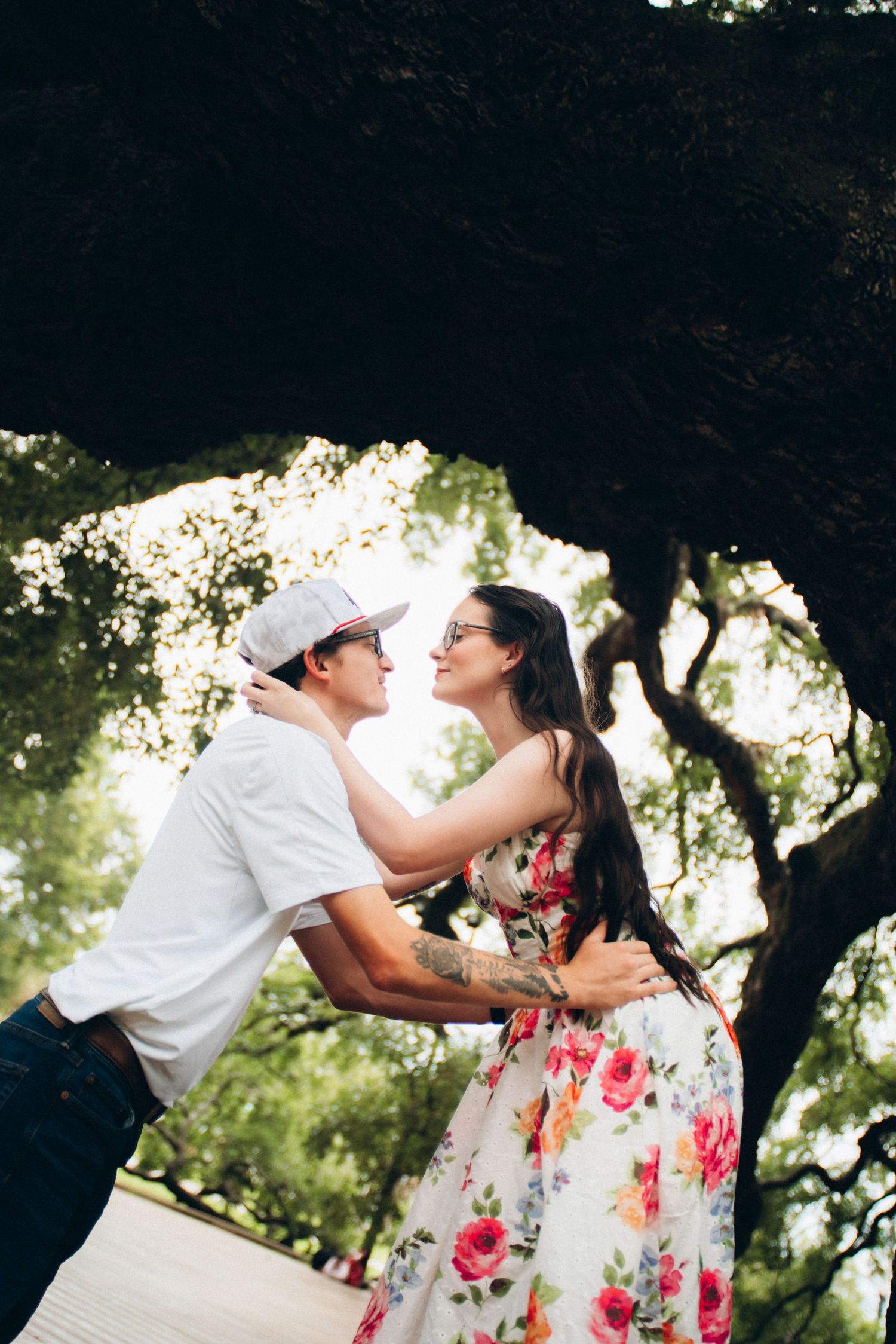 Romantic couple embracing and gazing at each other under large tree canopy, woman in floral dress, man in white cap.