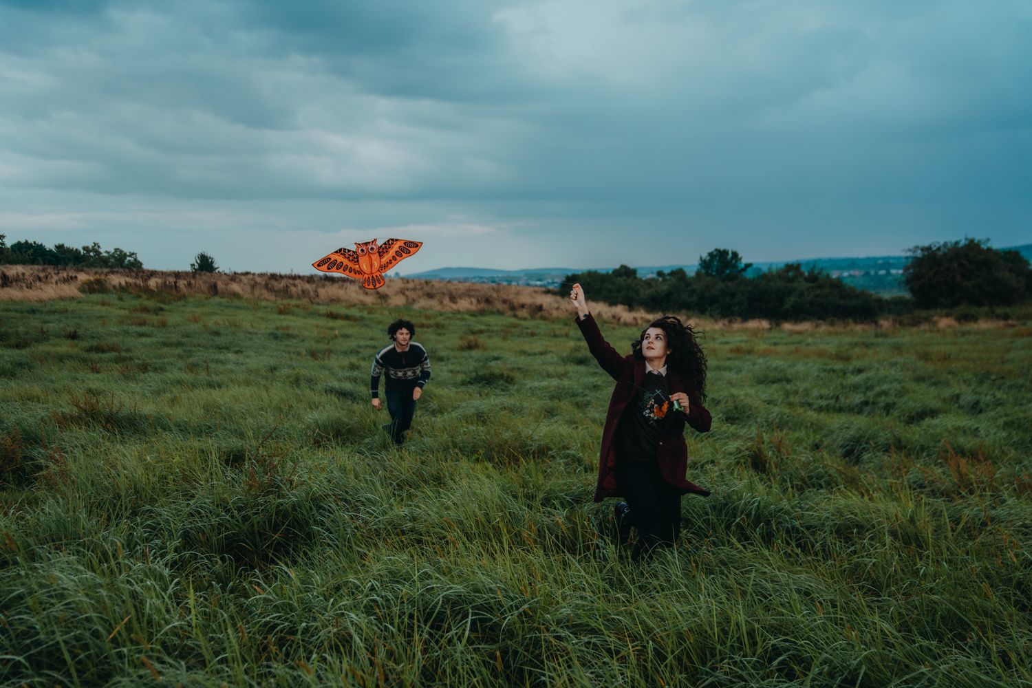Two people flying an orange kite in a vast green field under a dramatic cloudy sky.