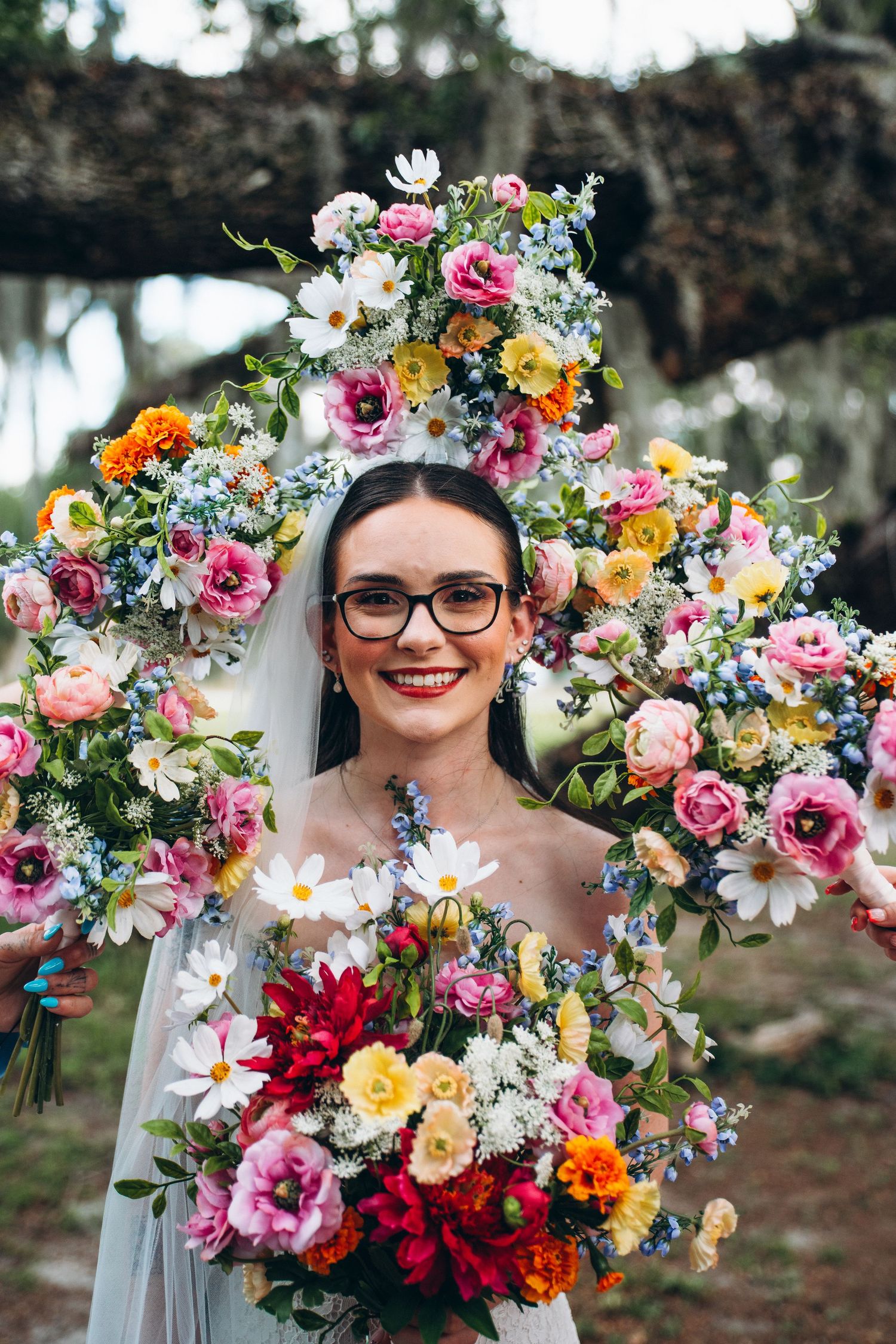 Smiling bride in glasses holding colorful floral bouquet, surrounded by vibrant flower arch with pink, white, and blue blooms.
