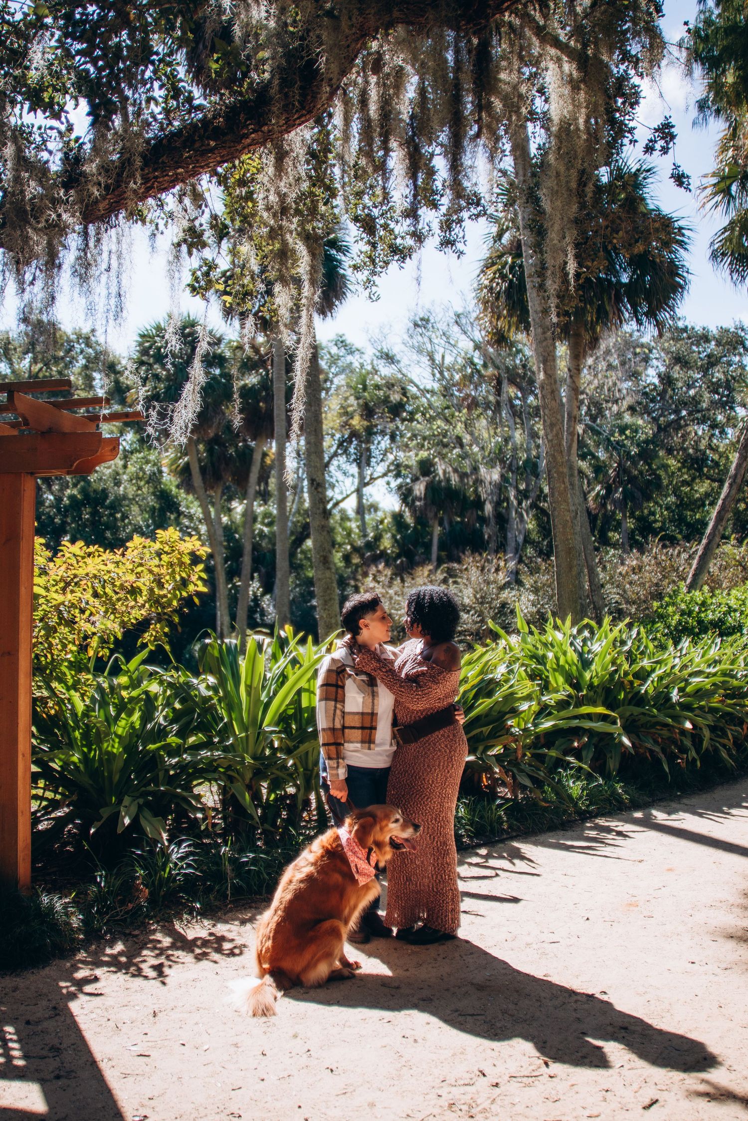A couple plays with their golden retriever in a tropical garden setting with hanging moss.