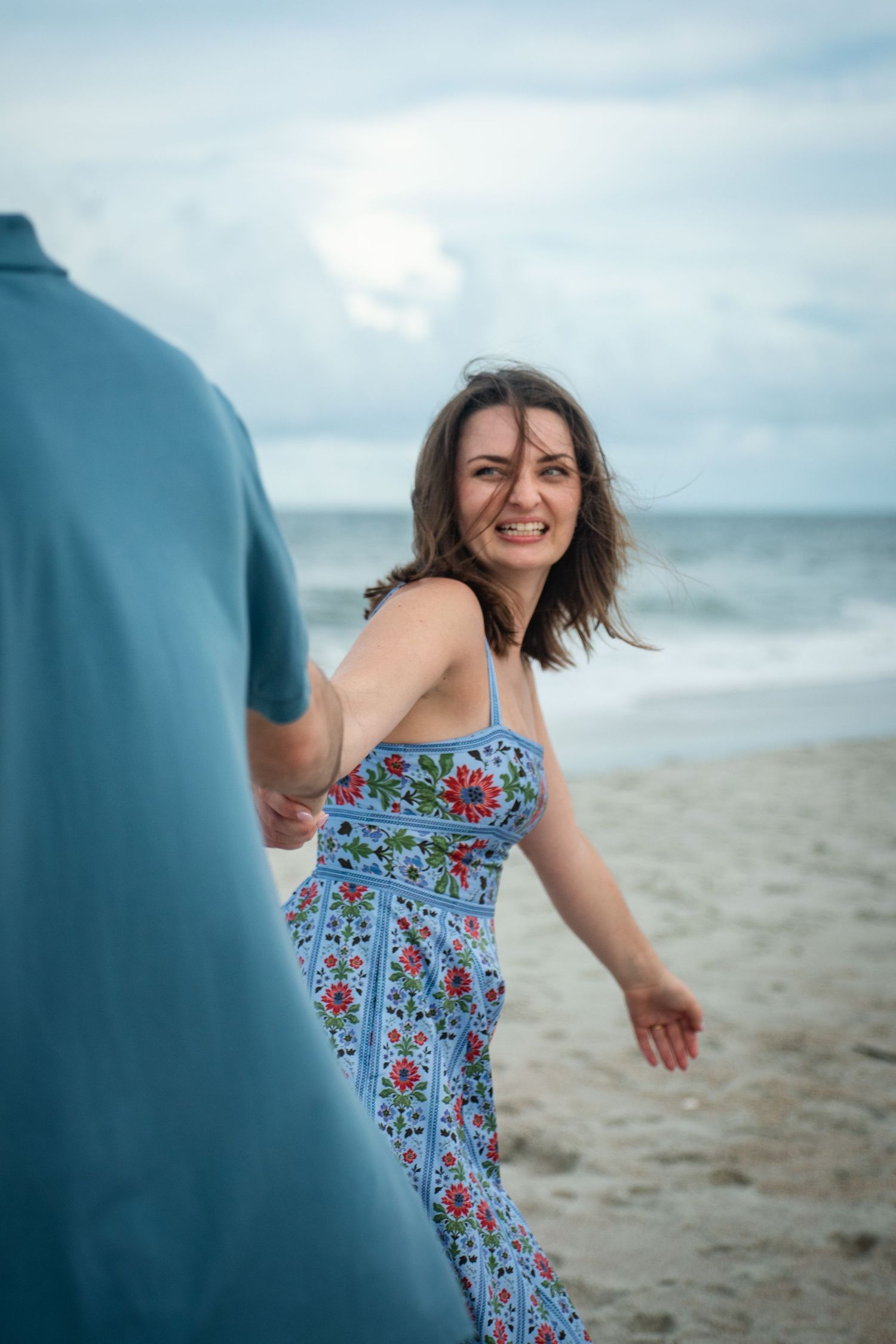A woman in a blue floral sundress smiles and dances joyfully on a cloudy beach.