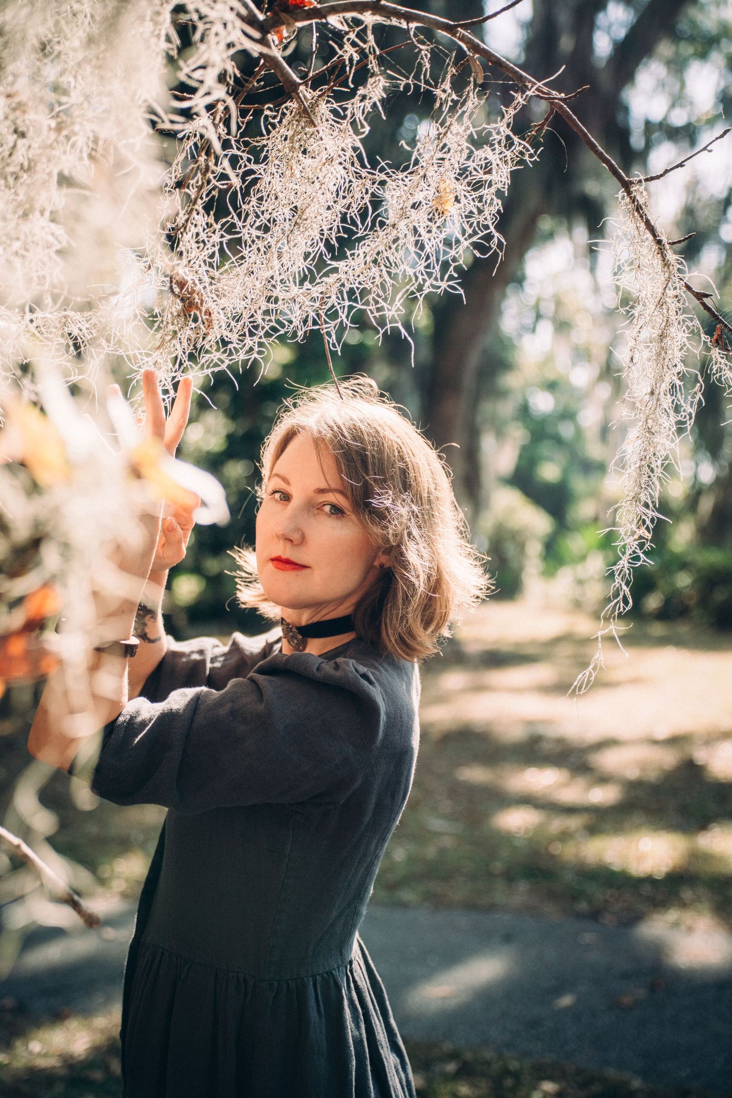 A person in black clothing reaches up to touch delicate branches against a sunlit forest backdrop.