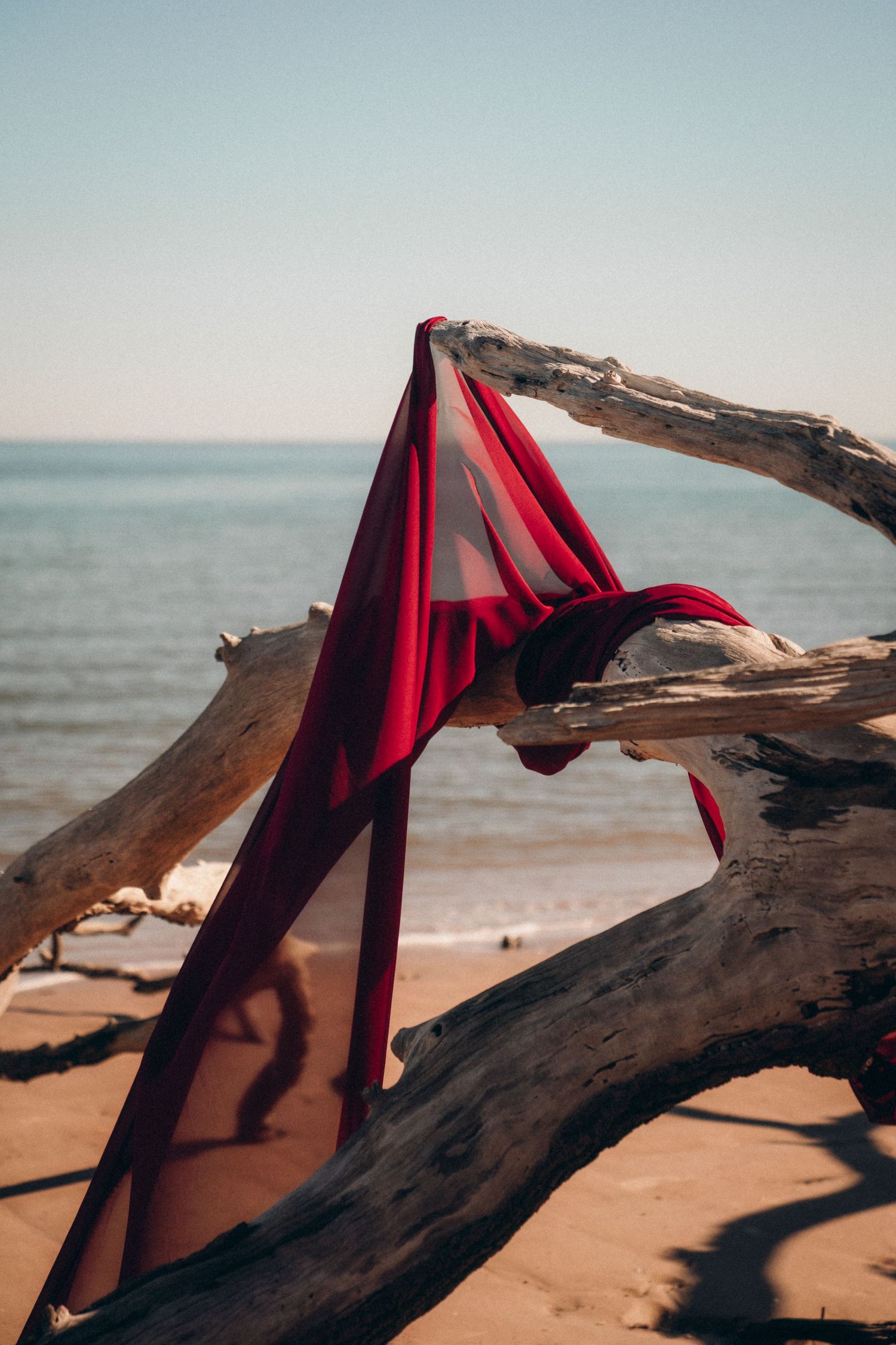 Red fabric draped over driftwood on a sandy beach with ocean waves in the background.