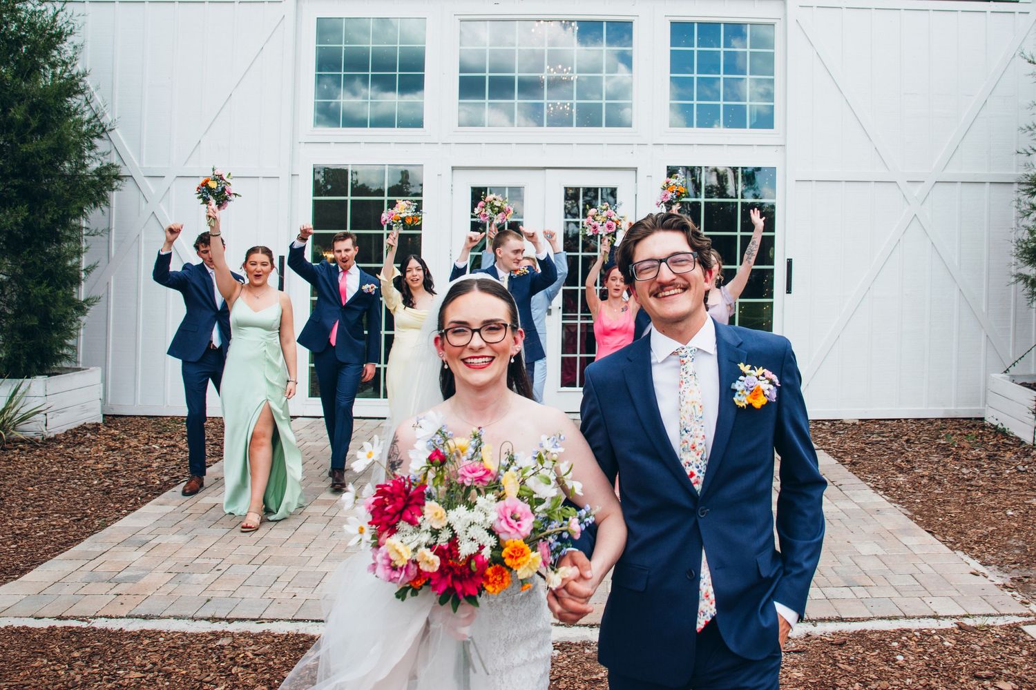 Smiling bride and groom hold hands in front of white barn doors as wedding party cheers behind them.