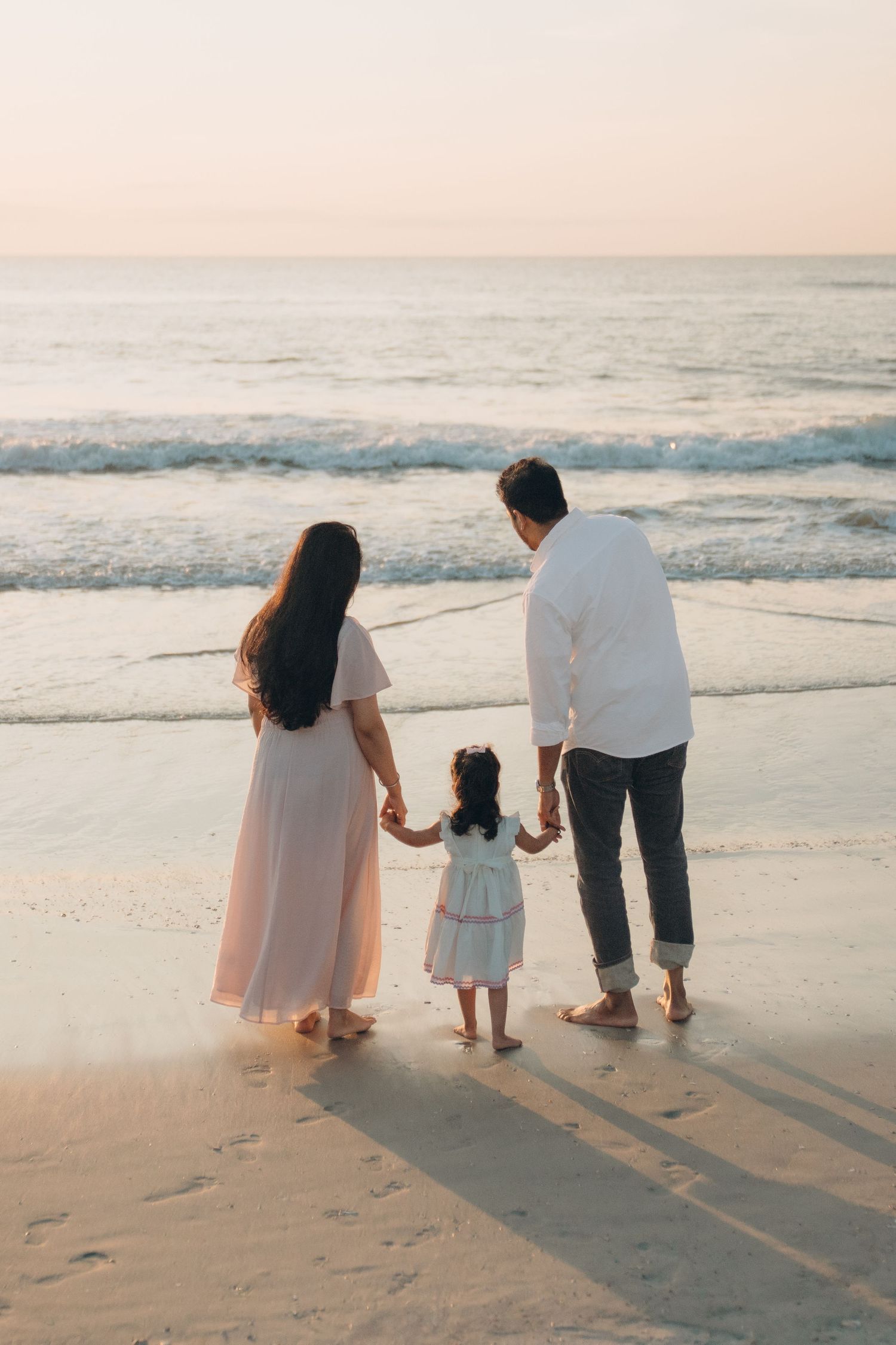 Family of three holding hands on a beach at sunset, parents and young daughter facing the ocean waves.