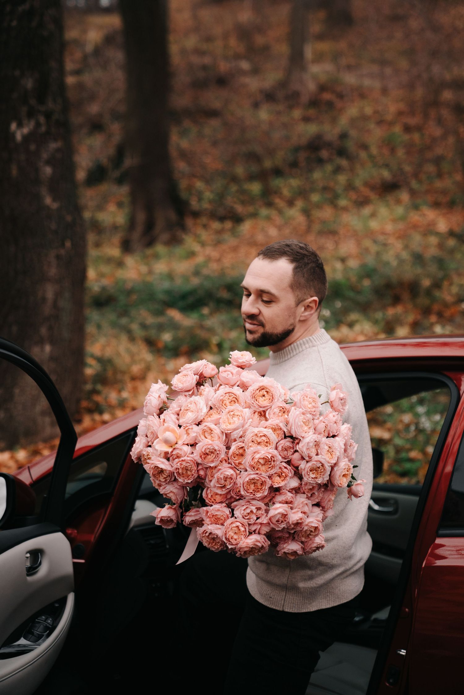 Bearded man in white sweater holding large bouquet of pink roses beside a red car.