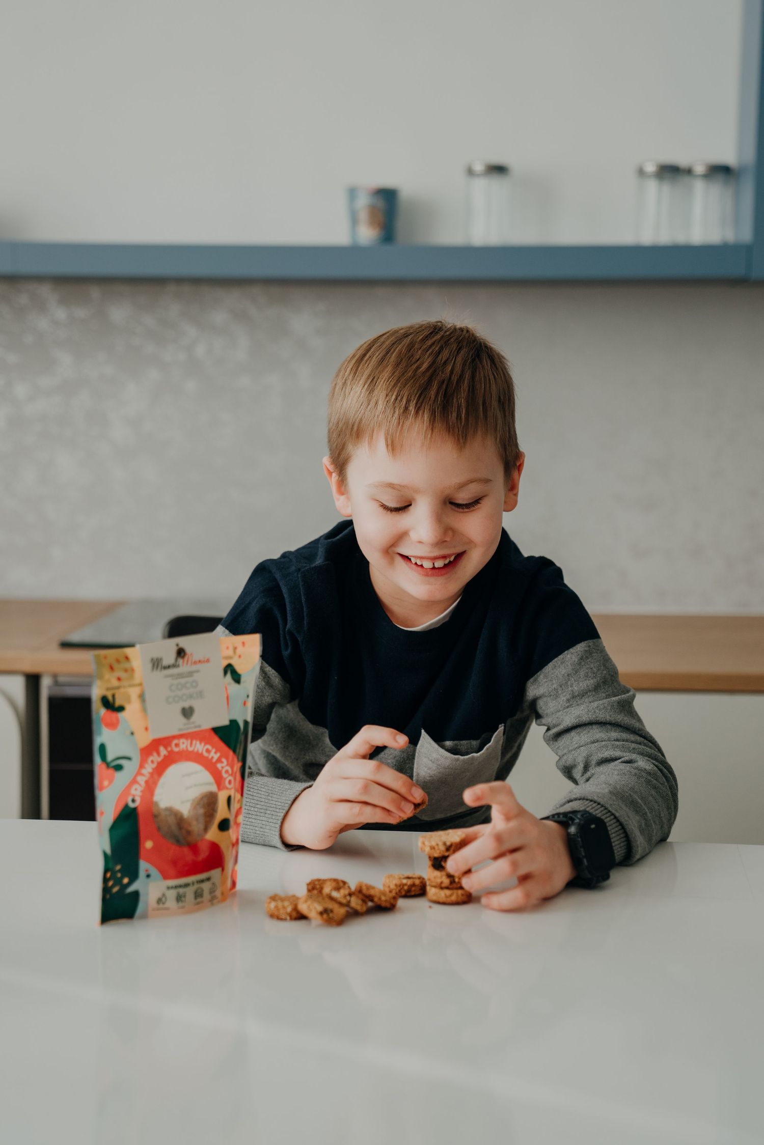 Happy young boy smiling at snack crackers on white table with colorful snack box in bright kitchen.