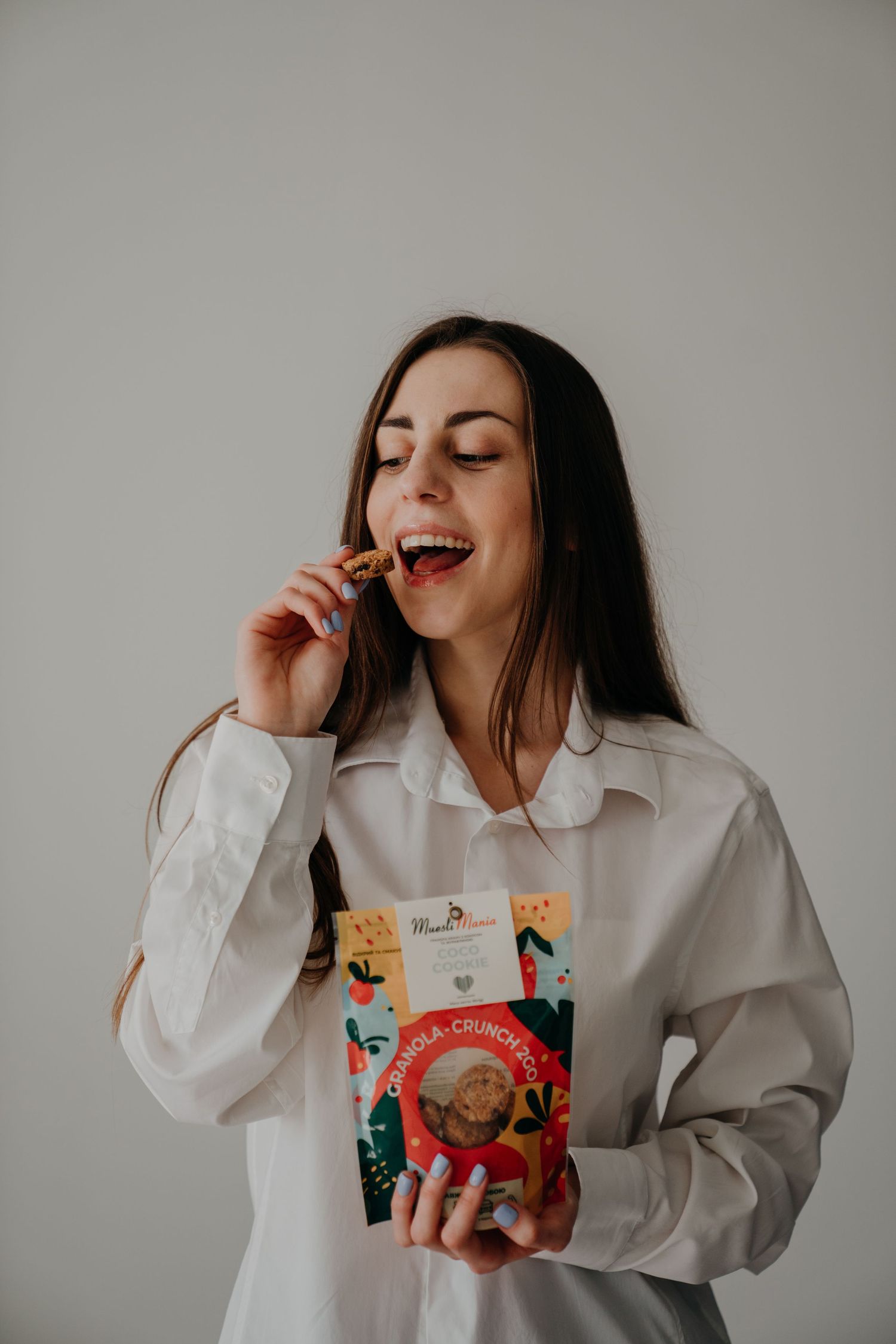 Smiling brunette woman in white shirt eating granola snack while holding MuesliMania Crunchco package.