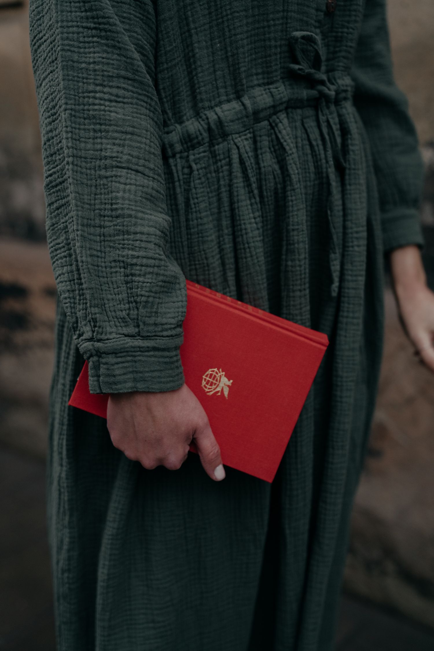 Person in green linen dress holding a red book with gold emblem outdoors.