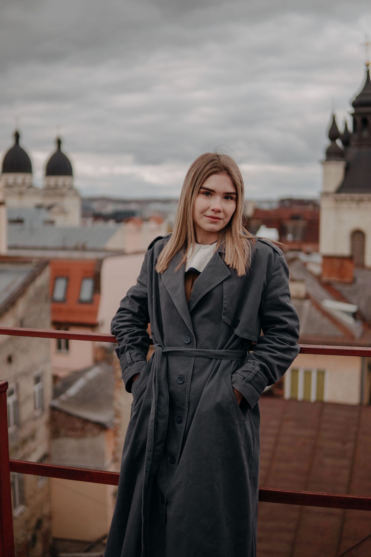 Blonde woman in gray trench coat stands on rooftop with European city architecture behind her.