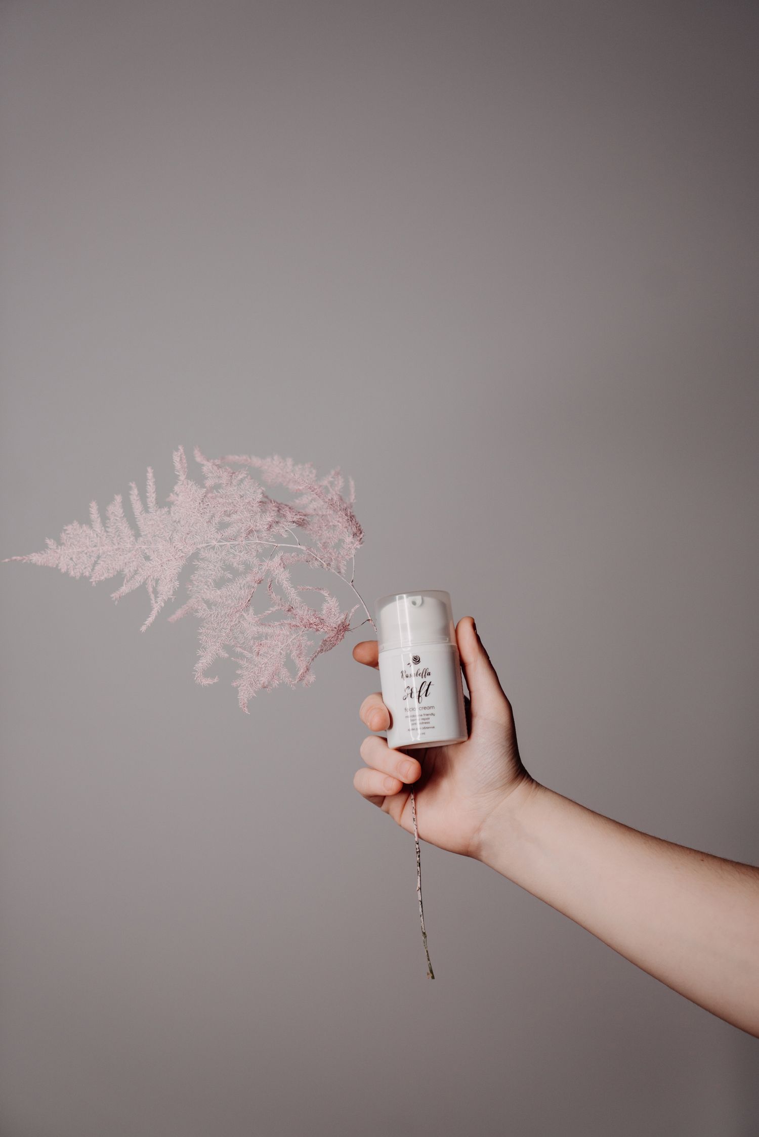 Hand holding a white Rosabella soft facial cream jar with pink dried fern decor.