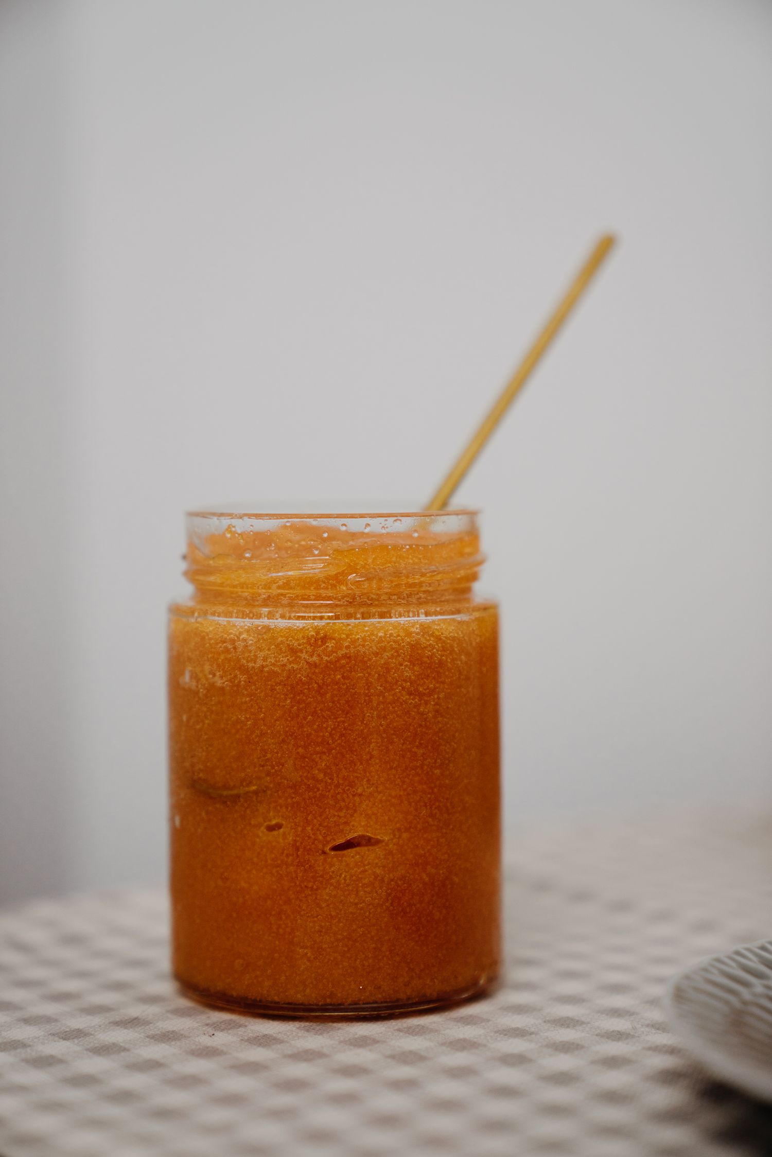 Glass jar of orange homemade jam or preserves with a wooden honey dipper stirrer on a checkered tablecloth.