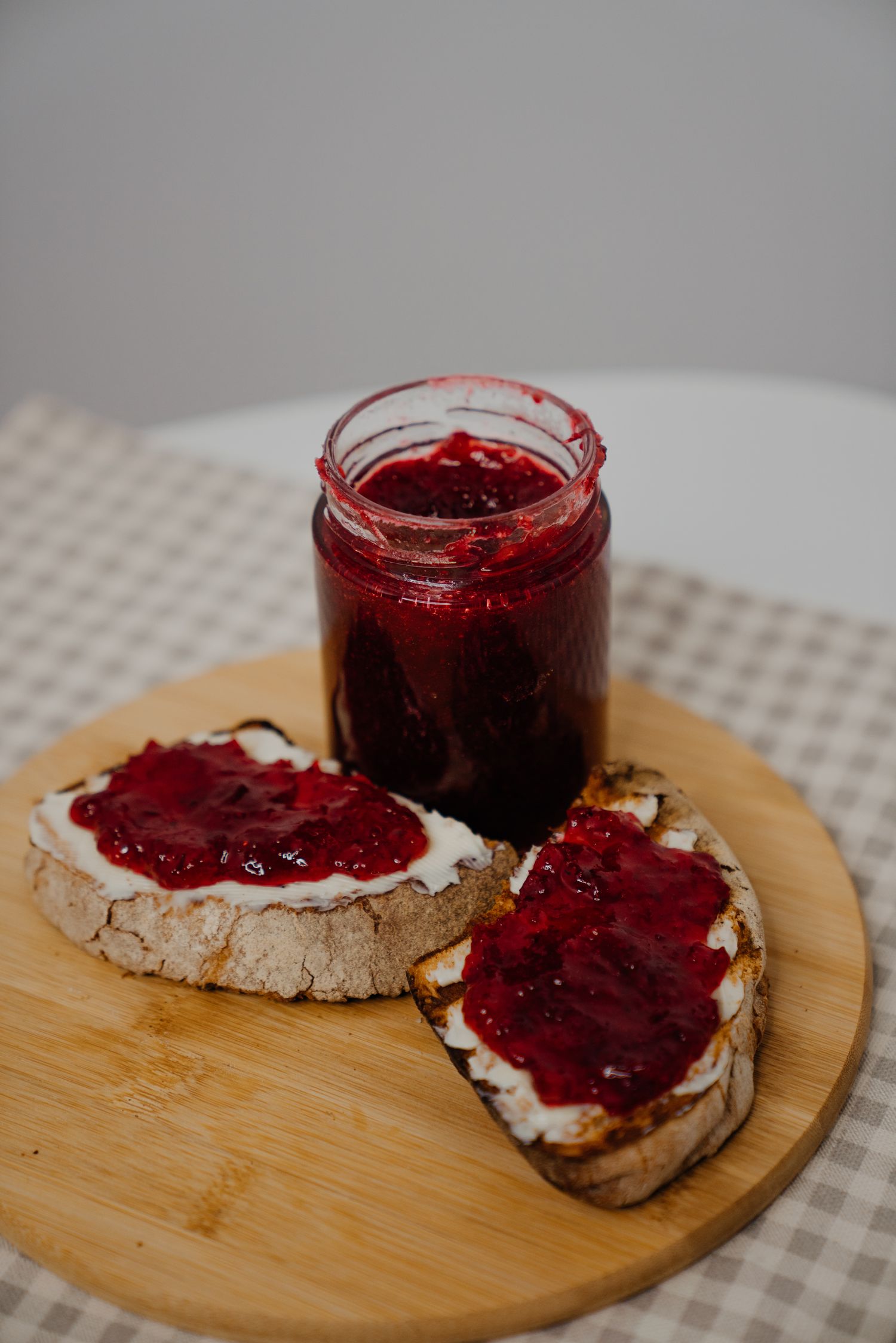 Open jar of homemade red berry jam on wooden board with two slices of bread spread with jam.