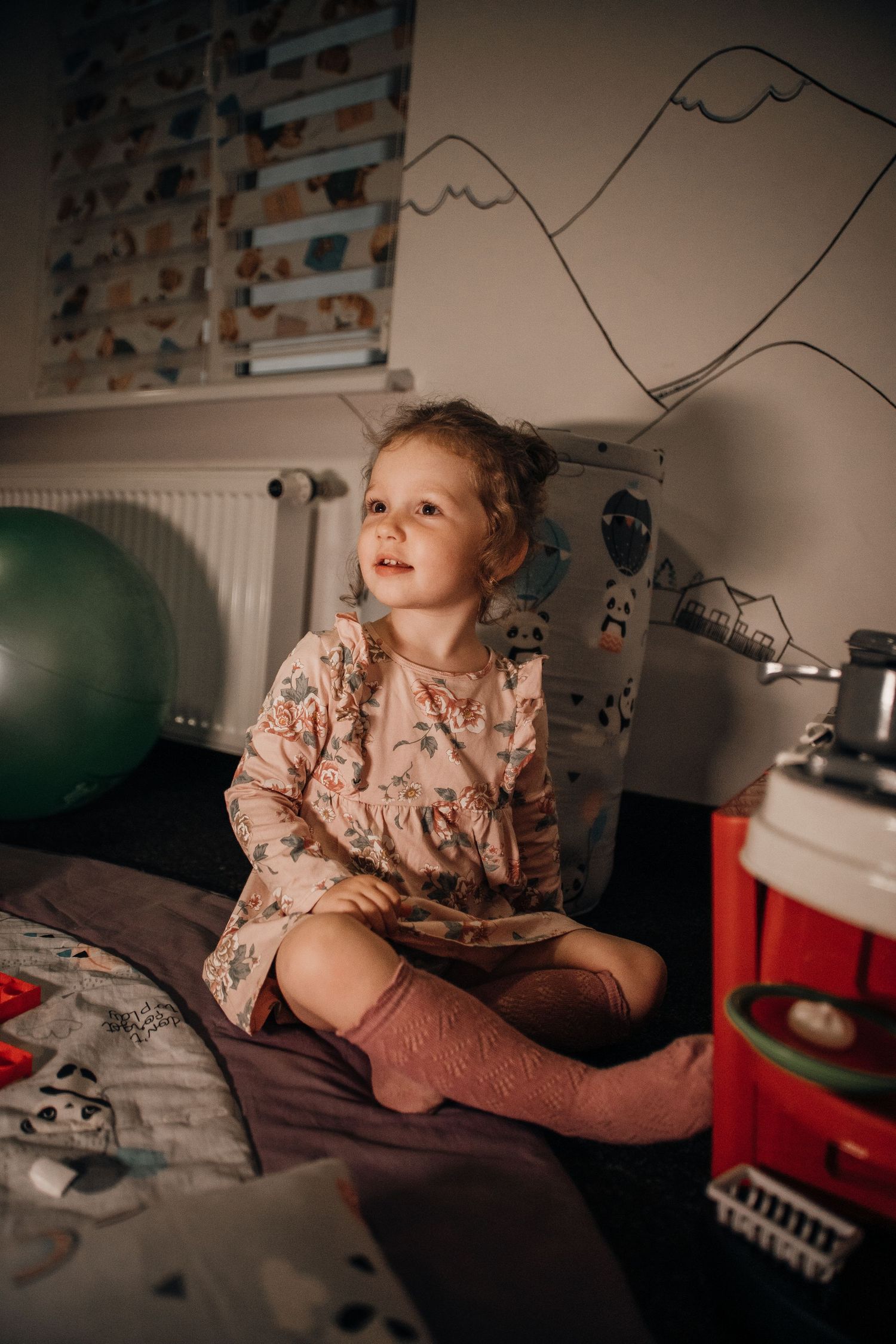 Little girl in floral dress sitting cross-legged on a bed in a decorated children's room.