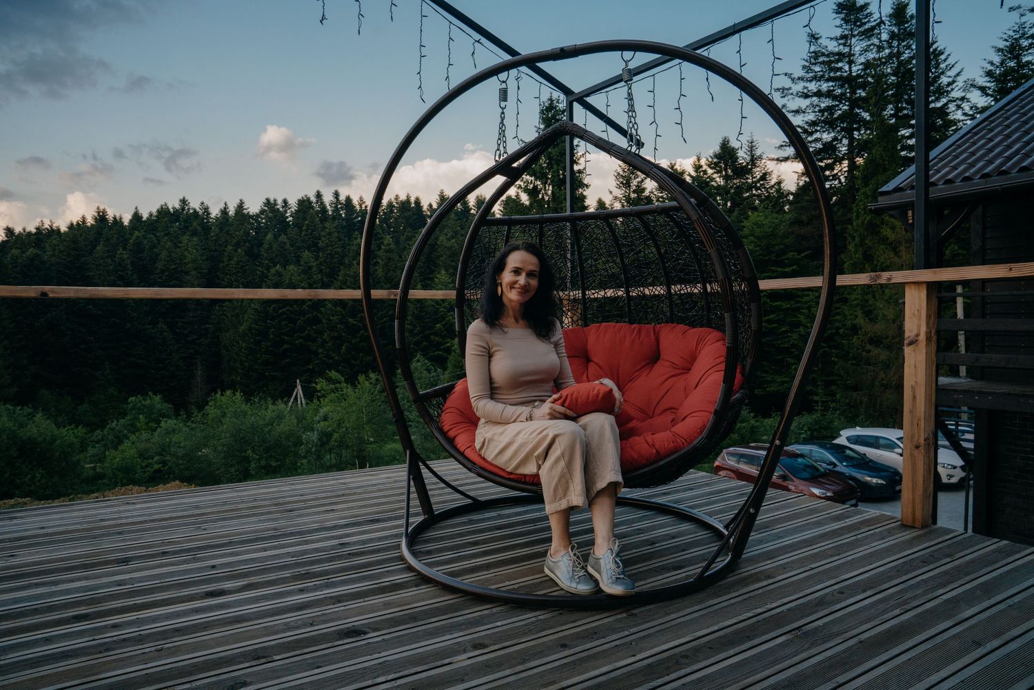 Woman relaxing in a hanging egg chair on a wooden deck surrounded by forest at dusk.