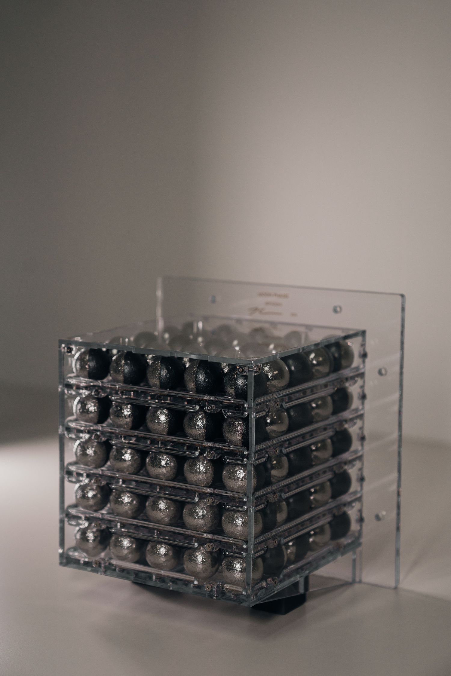 Clear acrylic cube display filled with rows of shiny metallic silver balls arranged in a grid pattern on a neutral background.