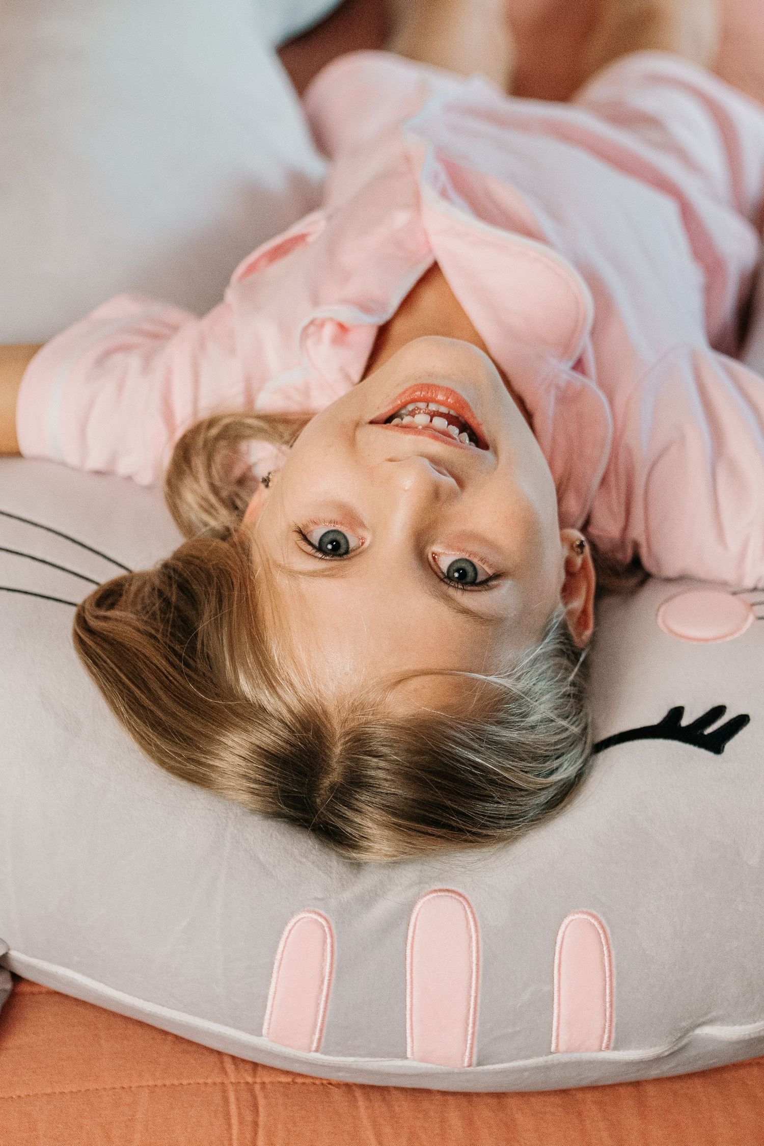 Young girl in pink pajamas lying upside down on a gray bunny-shaped pillow, smiling and laughing playfully.