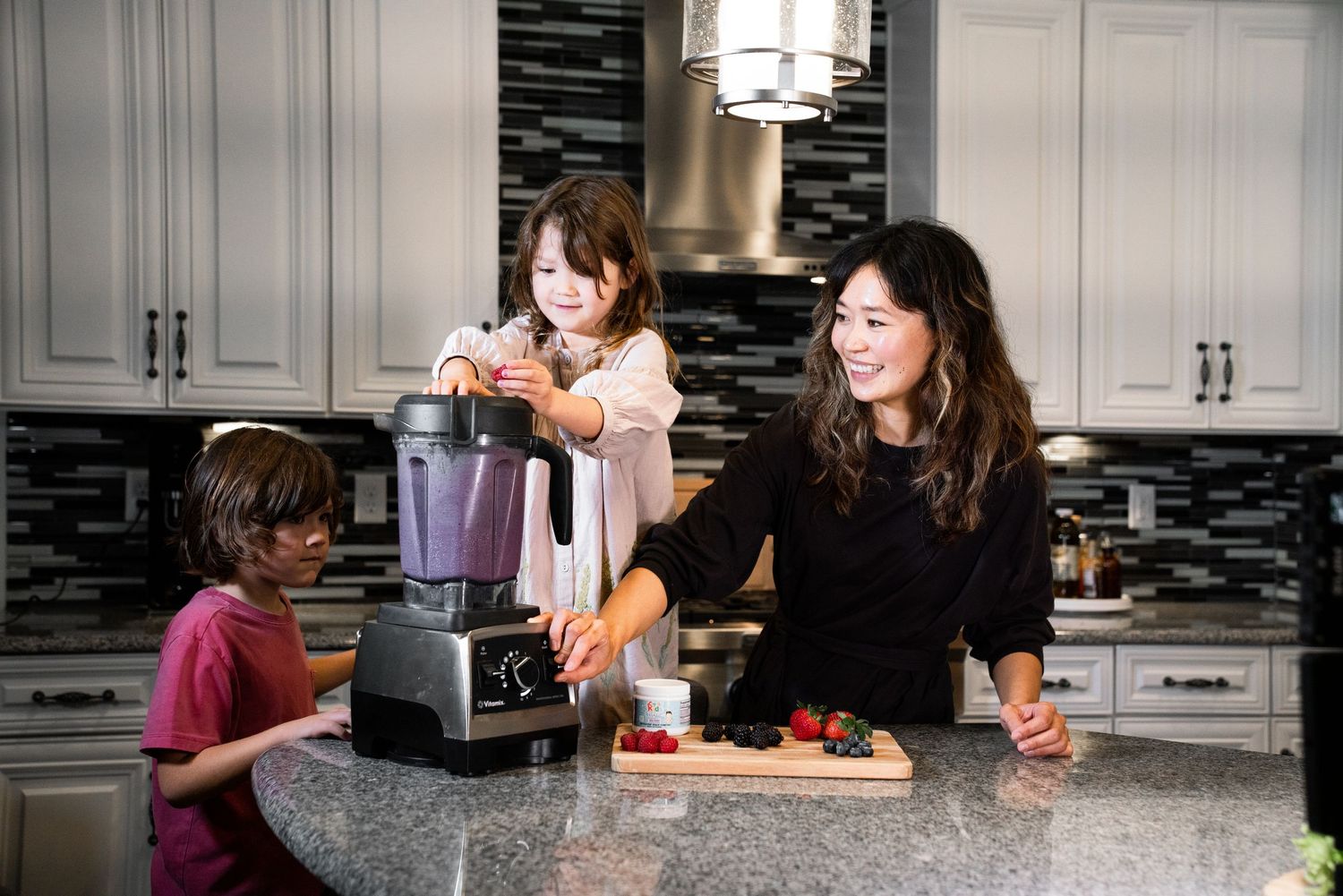 Woman and two children making smoothies with a blender and fresh strawberries in a modern kitchen.